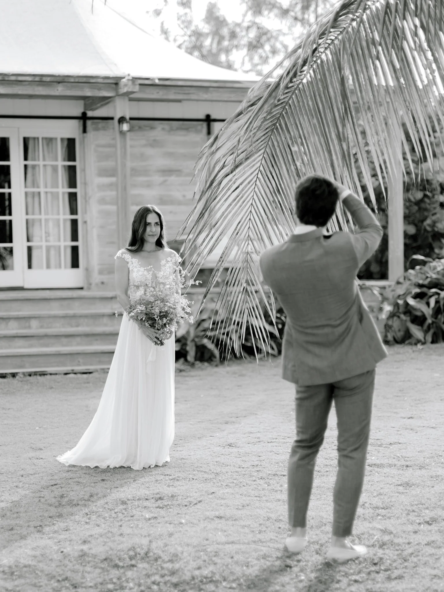 A woman in a wedding dress holding a bouquet stands outdoors in front of a wooden building, while a man in a suit, seen from the back, takes a photo of her behind a large palm leaf.