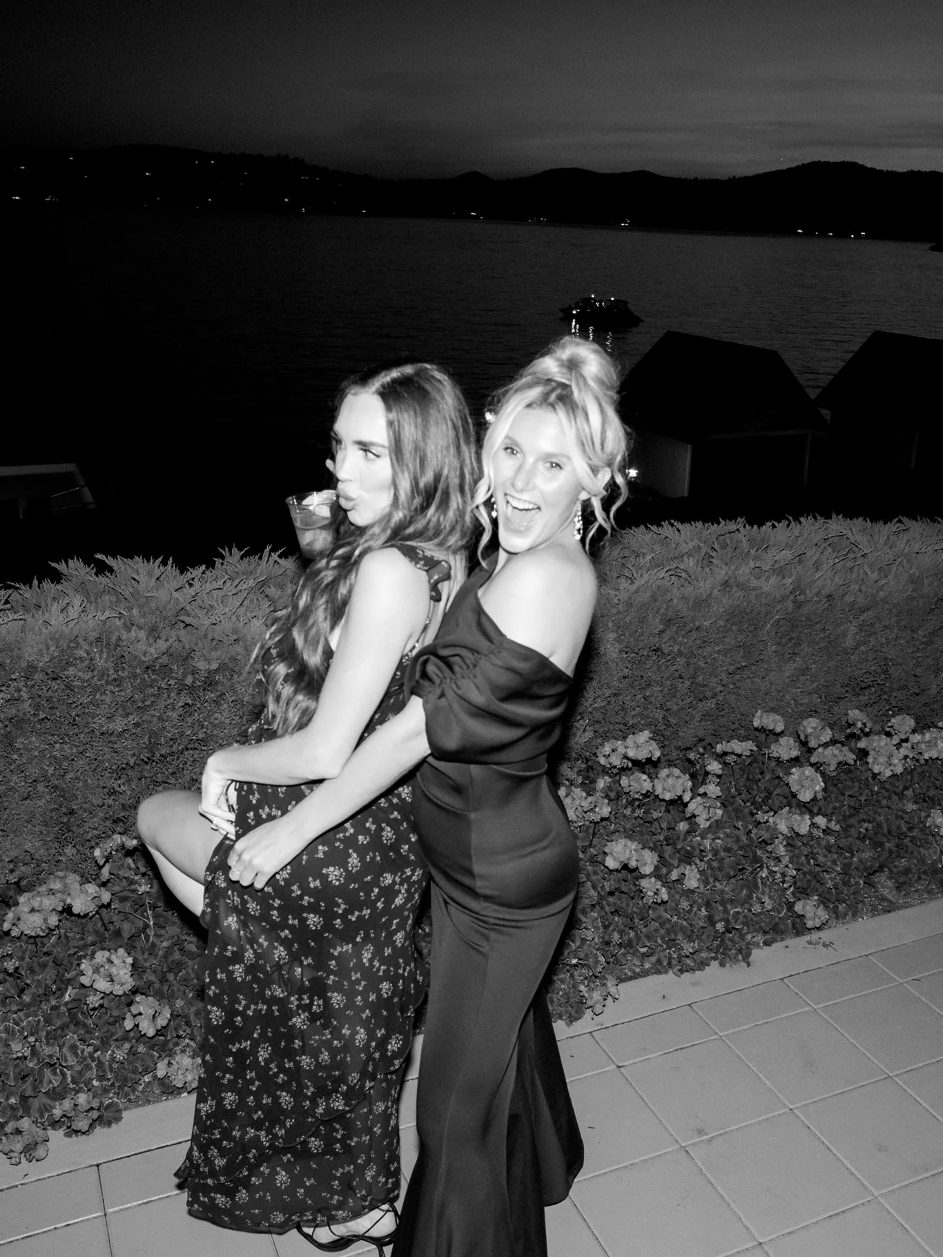 Two women in evening attire posing outdoors at night near a lake, with a boat and mountains in the background, smiling and having fun.