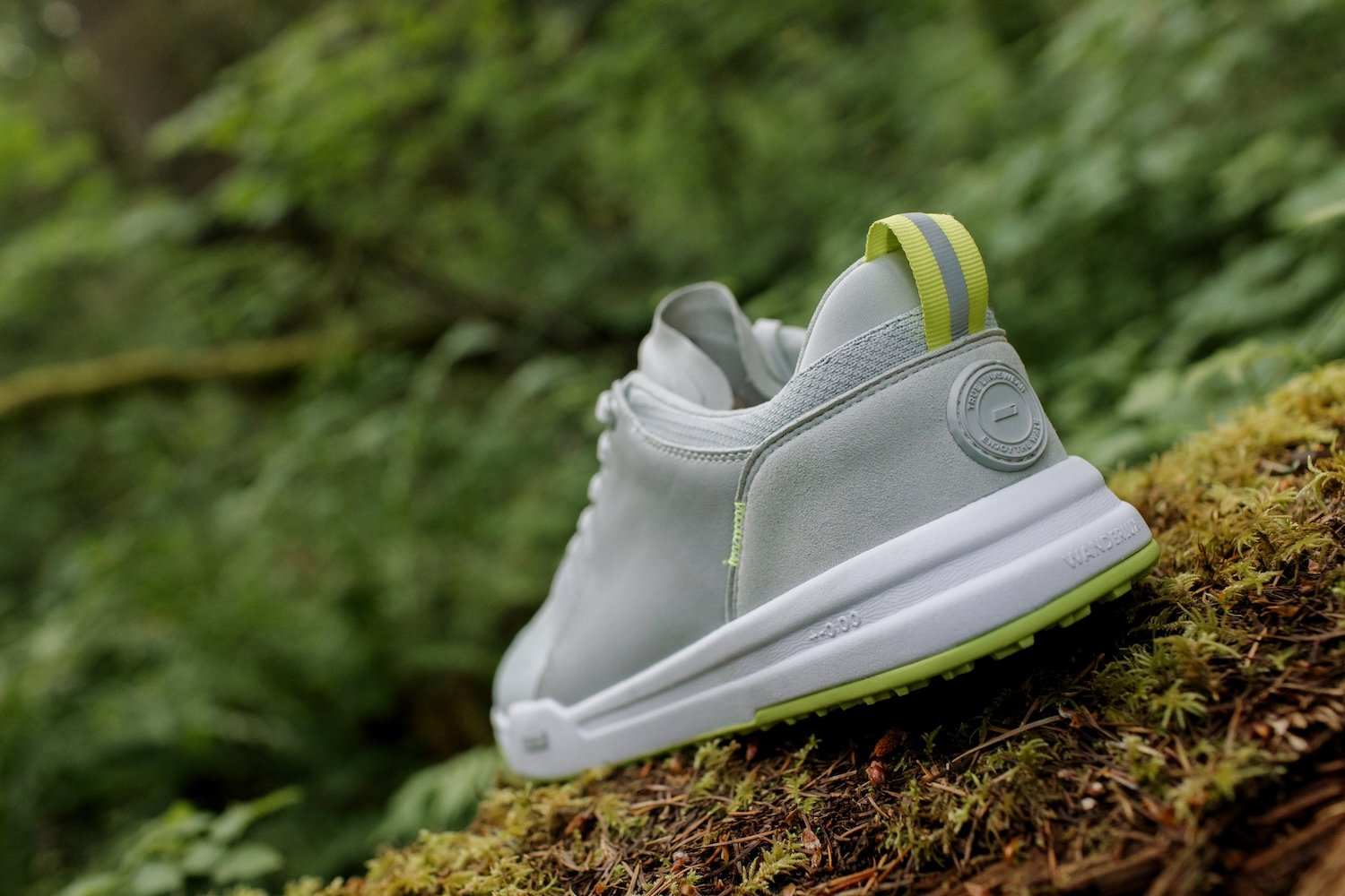 White athletic shoe with gray and neon yellow accents, placed outdoors on mossy ground with a blurred green forest background.