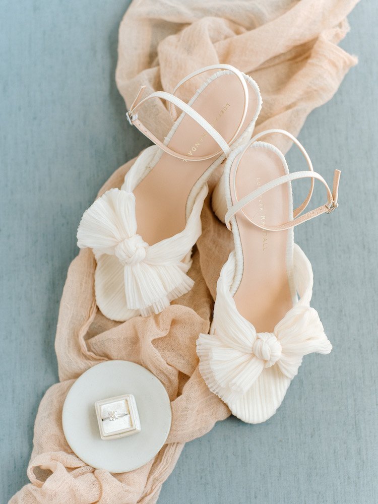 A pair of white high-heeled shoes with bows and crisscrossing ankle straps, set on beige and pink fabric. Nearby, a small ring box rests on a white plate.