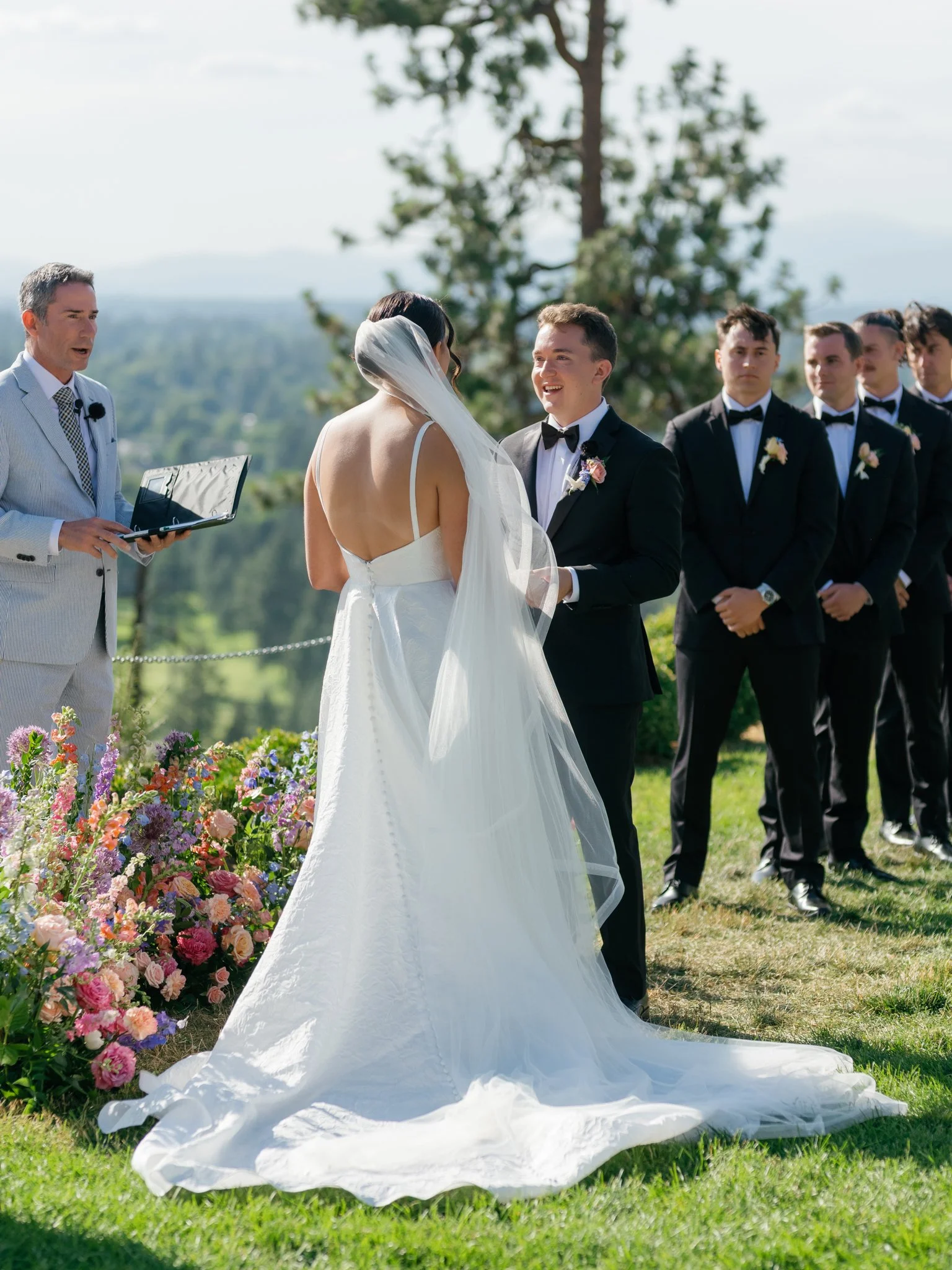 A wedding ceremony outdoors with the bride and groom exchanging vows, officiant speaking, and groomsmen standing in the background on a grassy area with trees and mountains in the distance.