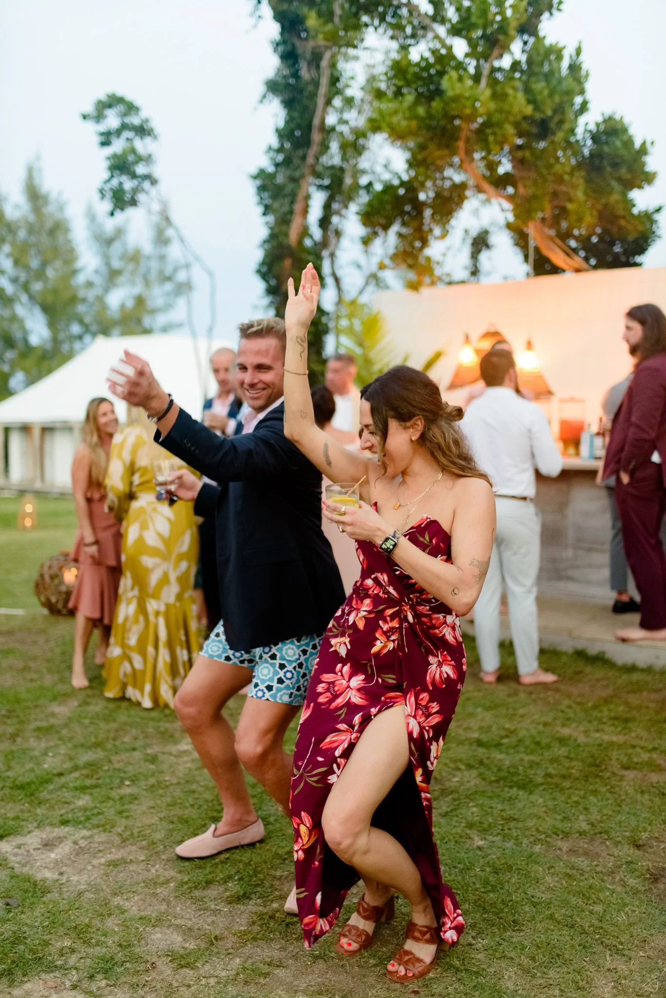 People dancing outdoors at a celebration or party, two women in floral dresses and a man in shorts and a blazer enjoying the moment.