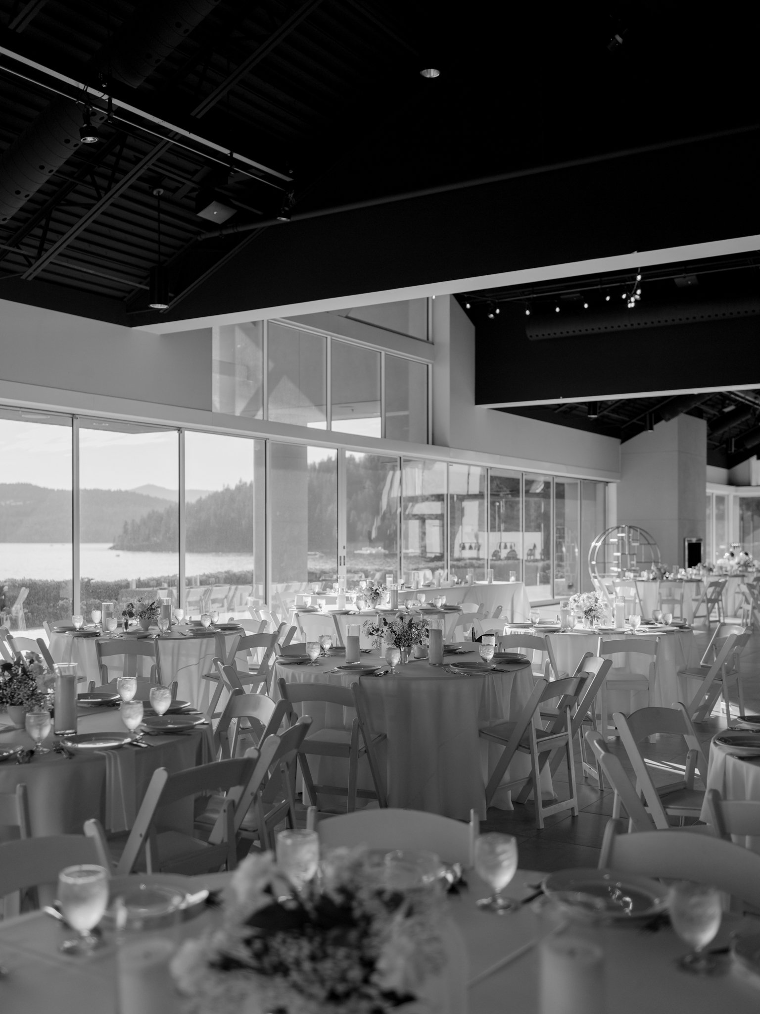 Indoor event space with round tables covered in white tablecloths, set with glasses, plates, and silverware, overlooking a scenic lake view through large glass windows at the Hagadone Event Center in Coeur d'Alene, Idaho.
