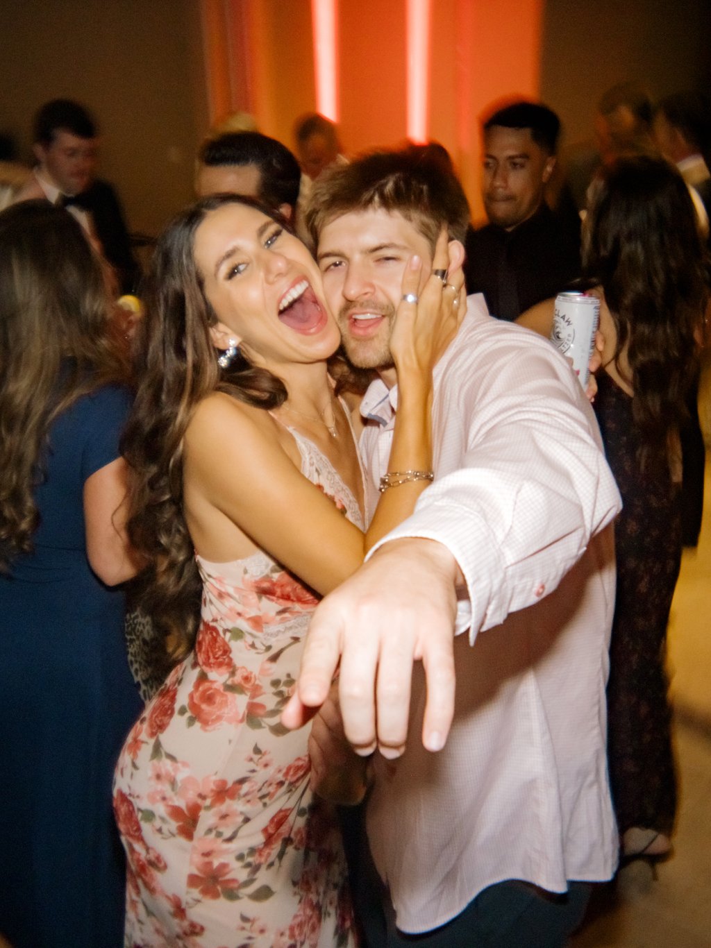 A woman and a man are dancing and posing together at a lively party, surrounded by other guests, with the woman smiling widely and the man making a playful face, holding a can of beer.
