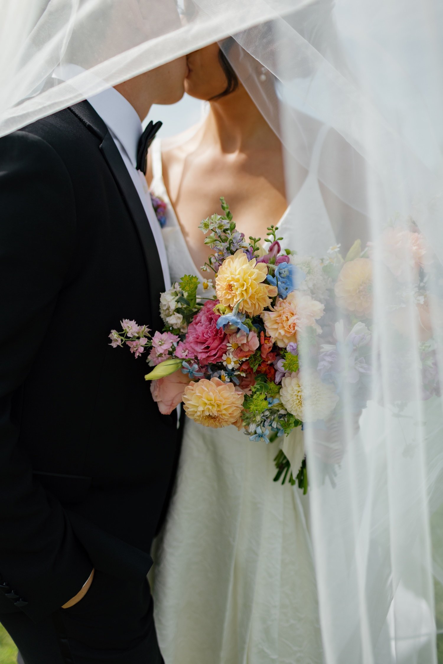 A bride and groom are close, kissing behind a veil at their wedding. The bride holds a colorful bouquet of flowers including roses, daisies, and other blooms.