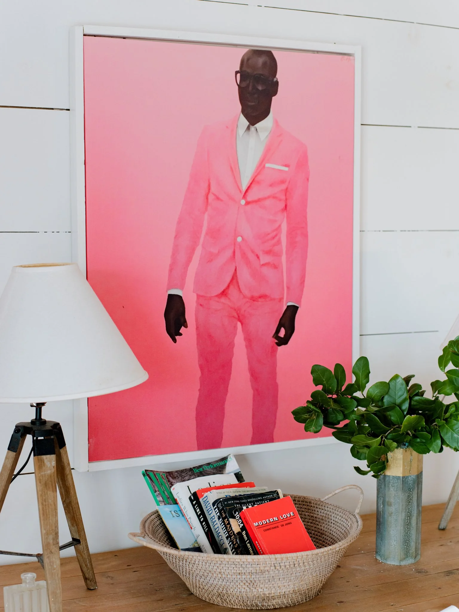 A large framed photo of a man wearing a pink suit, white shirt, dark sunglasses, and black gloves, standing against a pink background. There is a white wall with horizontal paneling behind the photo, a white lampshade on a wooden tripod lamp to the left, a woven basket with books in front of the photo, and a potted plant with green leaves in a marble and wood vase to the right.