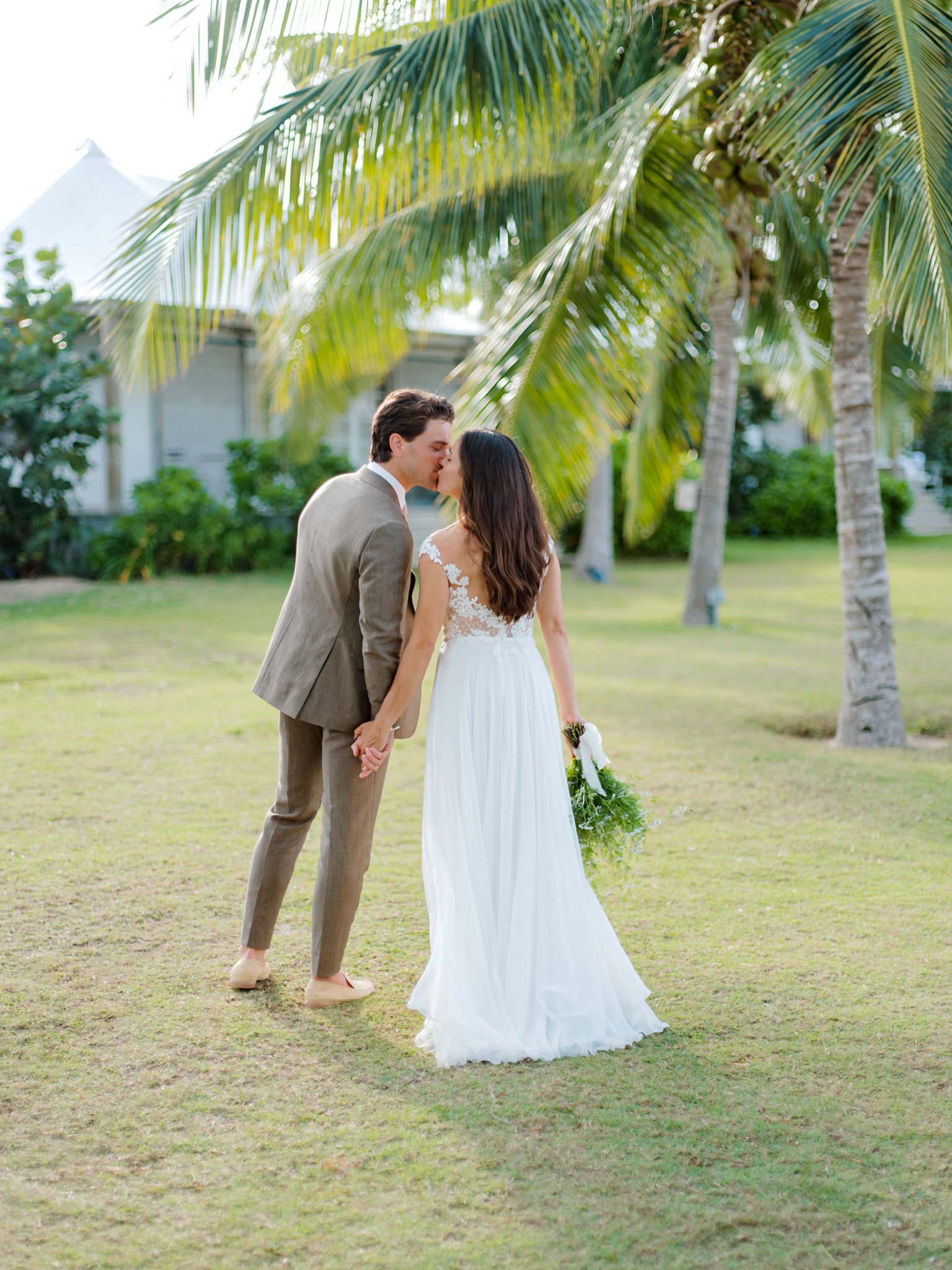 A newlywed couple sharing a kiss in an outdoor setting with palm trees and green foliage, the bride is in a white wedding dress holding a bouquet, and the groom is in a beige suit at The Other Side Hotel Eleuthera during a luxury Bahamas Wedding.