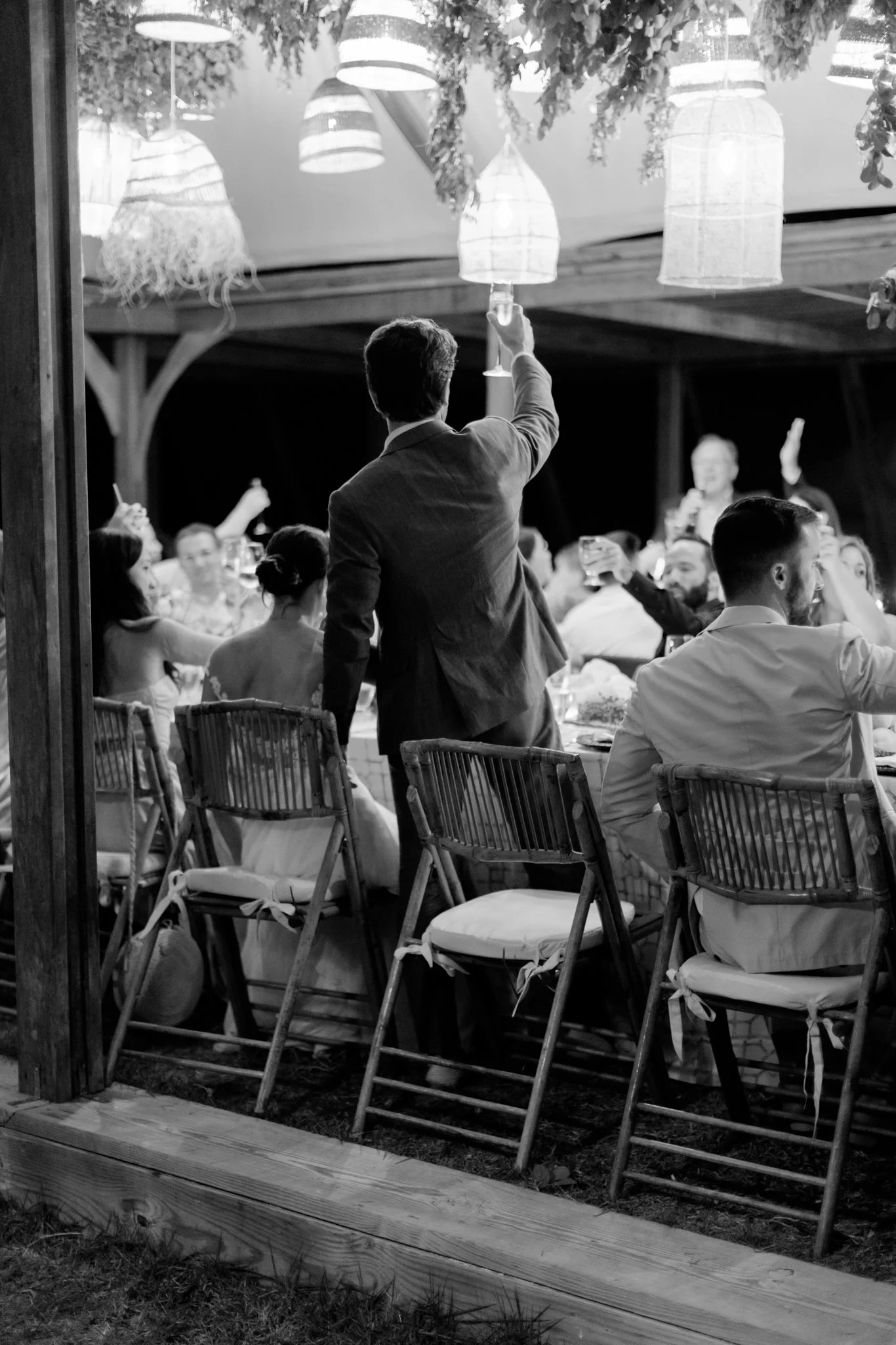 Black and white photo of a man in a suit raising a glass at a wedding reception with guests sitting at tables, hanging lanterns, and string lights overhead.
