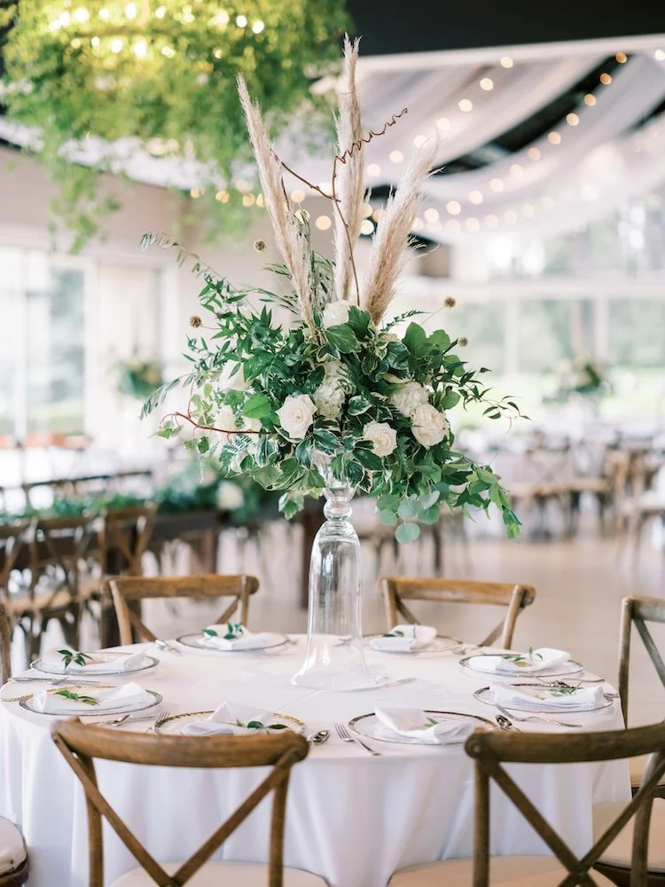Elegant banquet table with a large floral centerpiece featuring white roses, greenery, and pampas grass, surrounded by wooden chairs, set with plates, napkins, and cutlery, in a decorated reception hall with string lights and draped ceiling