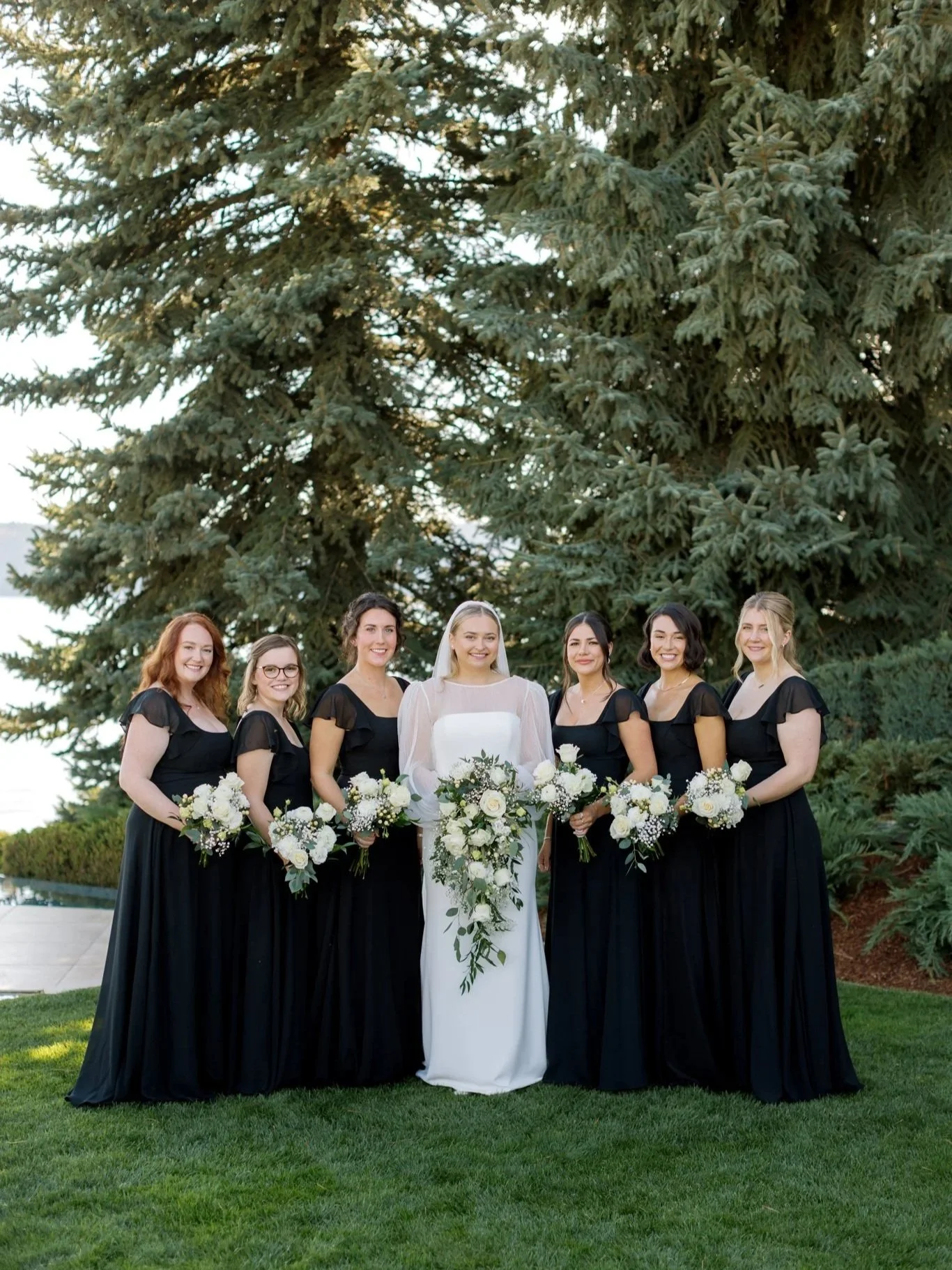 Bride standing with her bridesmaids in black dresses holding white bouquets on the lawn at the Hagadone Event Center overlooking Lake Coeur d’Alene.