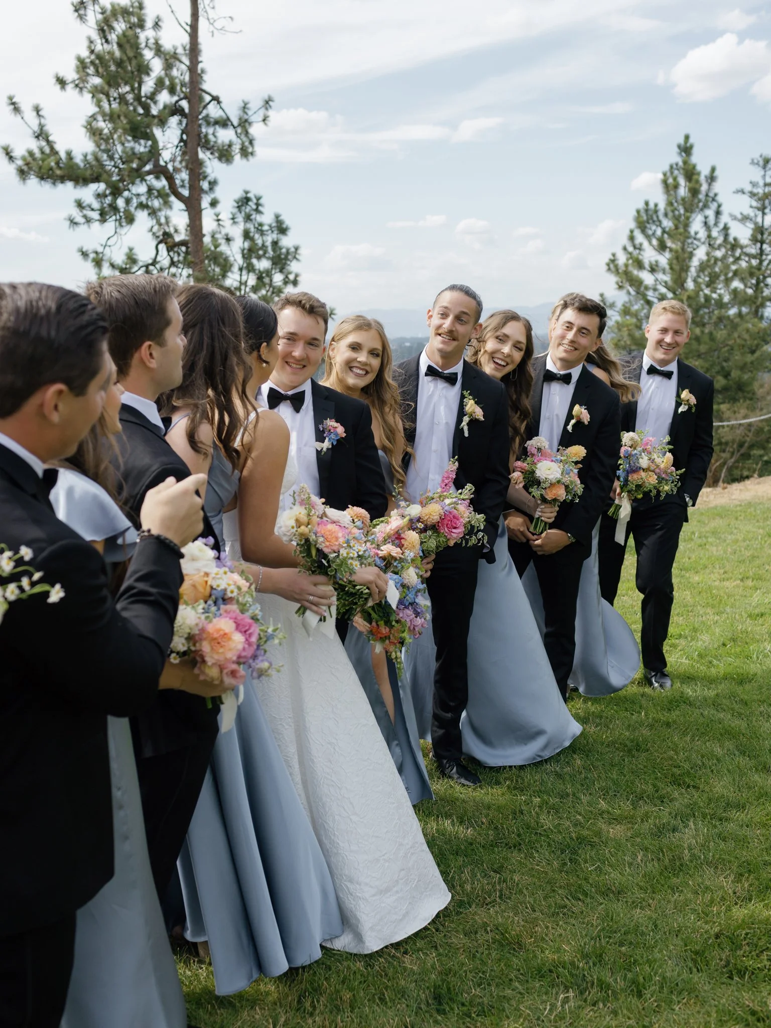 A wedding party standing outdoors on grass, with trees and mountains in the background, holding bouquets, and smiling in North Idaho, Coeur d'Alene.
