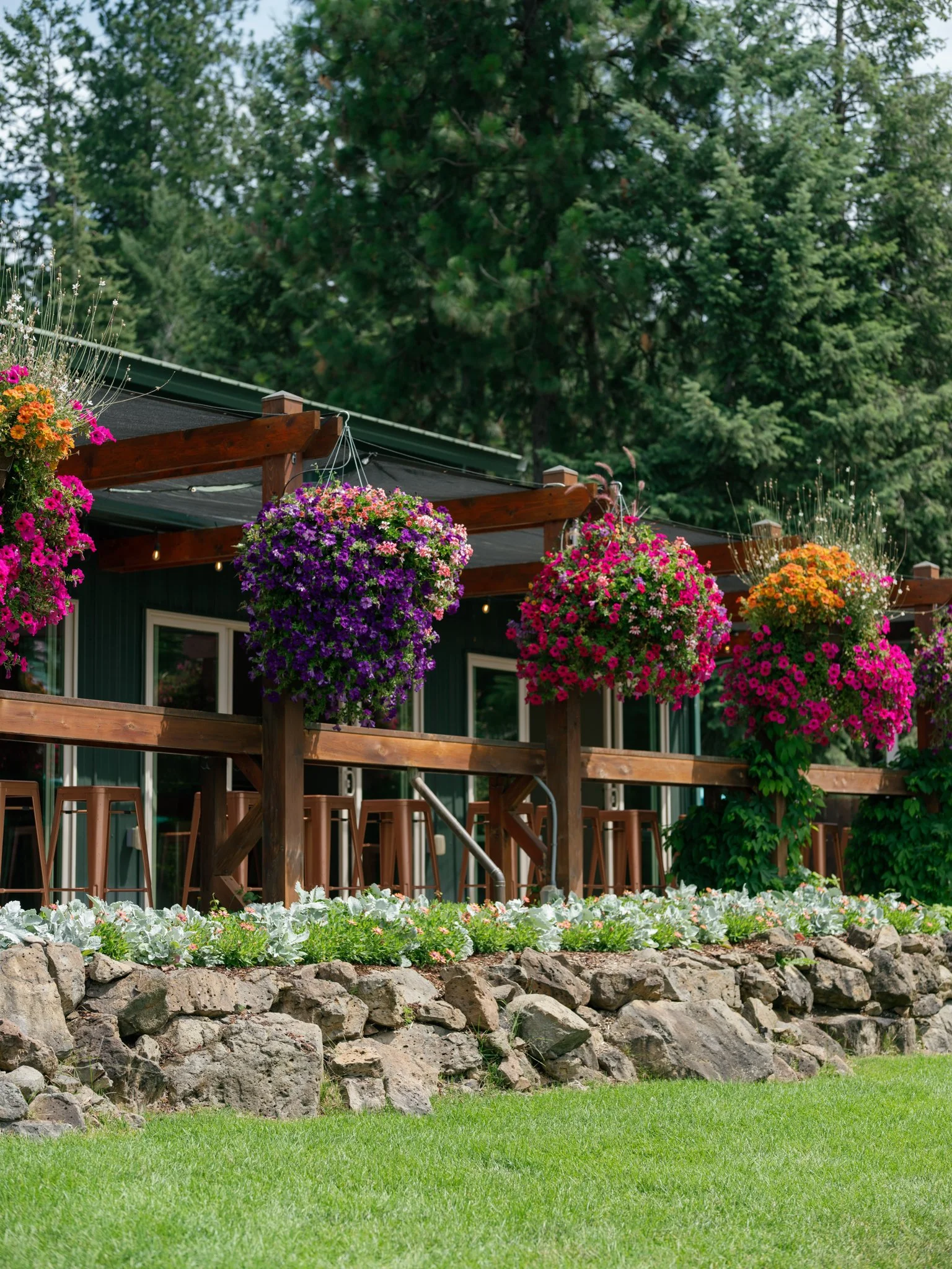 A wooden outdoor patio with hanging flower baskets in purple, pink, and orange, surrounded by a rock garden and greenery, with a forest in the background at Elk Point wedding above Lake Coeur d’Alene.