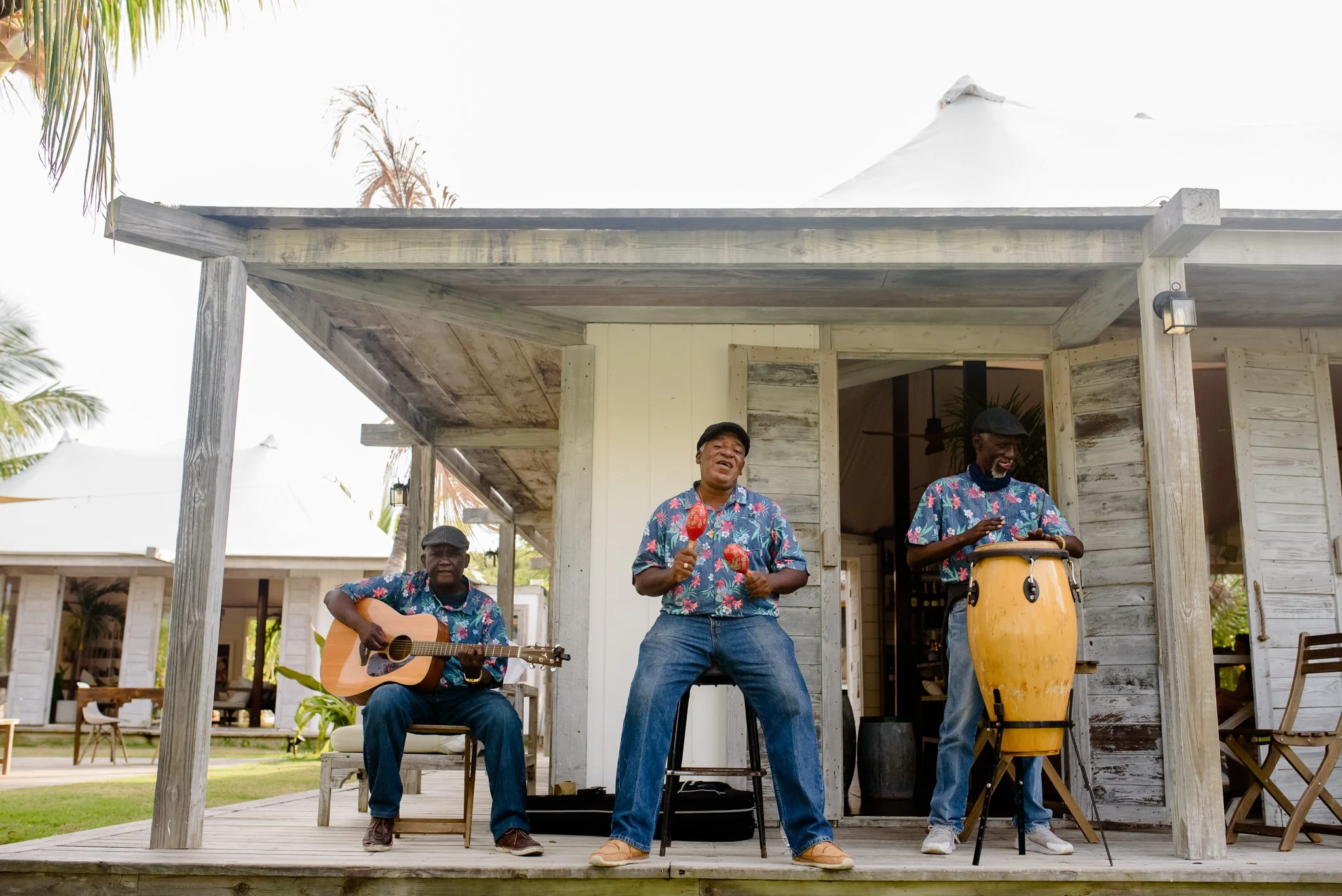 Three men performing music and singing on a wooden porch, one playing guitar, one holding maracas, and one playing a percussion instrument, all wearing matching floral shirts.