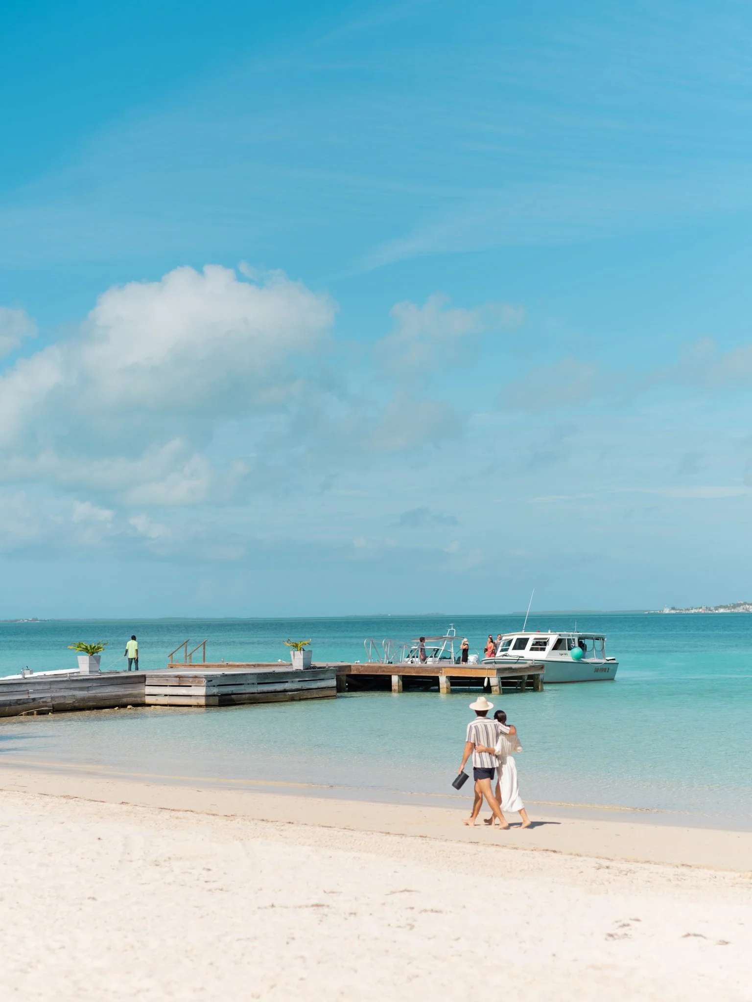 People walking along a beach with a pier and boat in the background, under a partly cloudy sky at The Other Side Hotel during their destination wedding in Eleuthera, Bahamas.