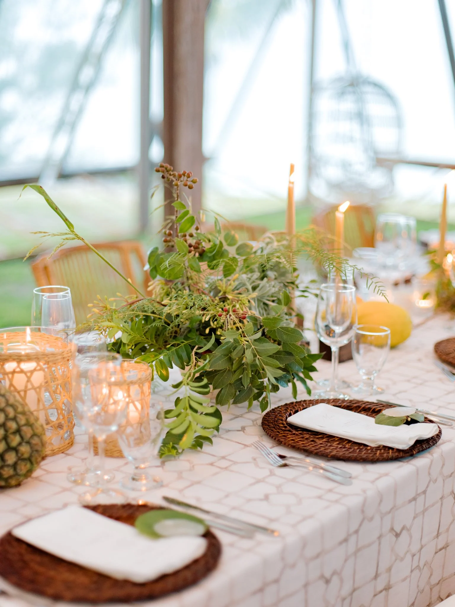 Decorative table setting with greenery centerpiece, candles, glasses, woven placemats, fork and knife, and napkins in a bright, airy tented space.