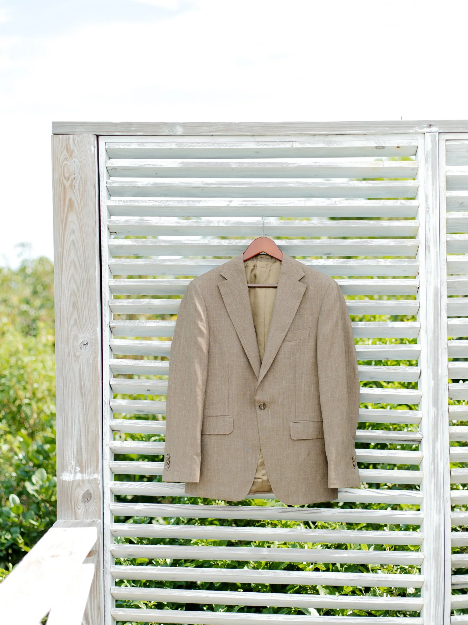 A beige blazer hanging on a pink hanger on a white wooden slatted panel outdoors with green foliage visible in the background.