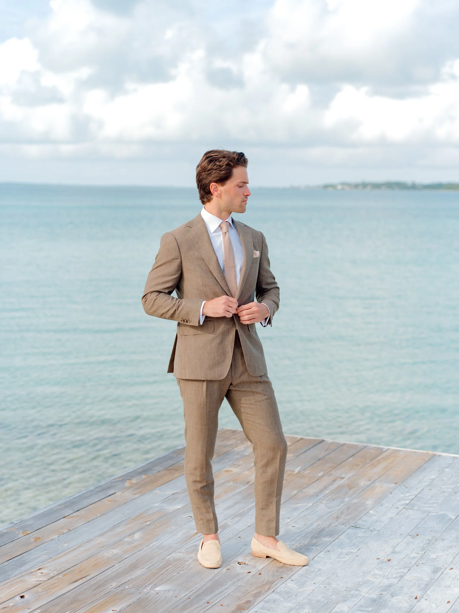A man in a tan suit and light-colored shoes stands on a wooden pier by the water with an ocean and cloudy sky in the background at The Other Side Hotel Eleuthera during a luxury Bahamas destination wedding.