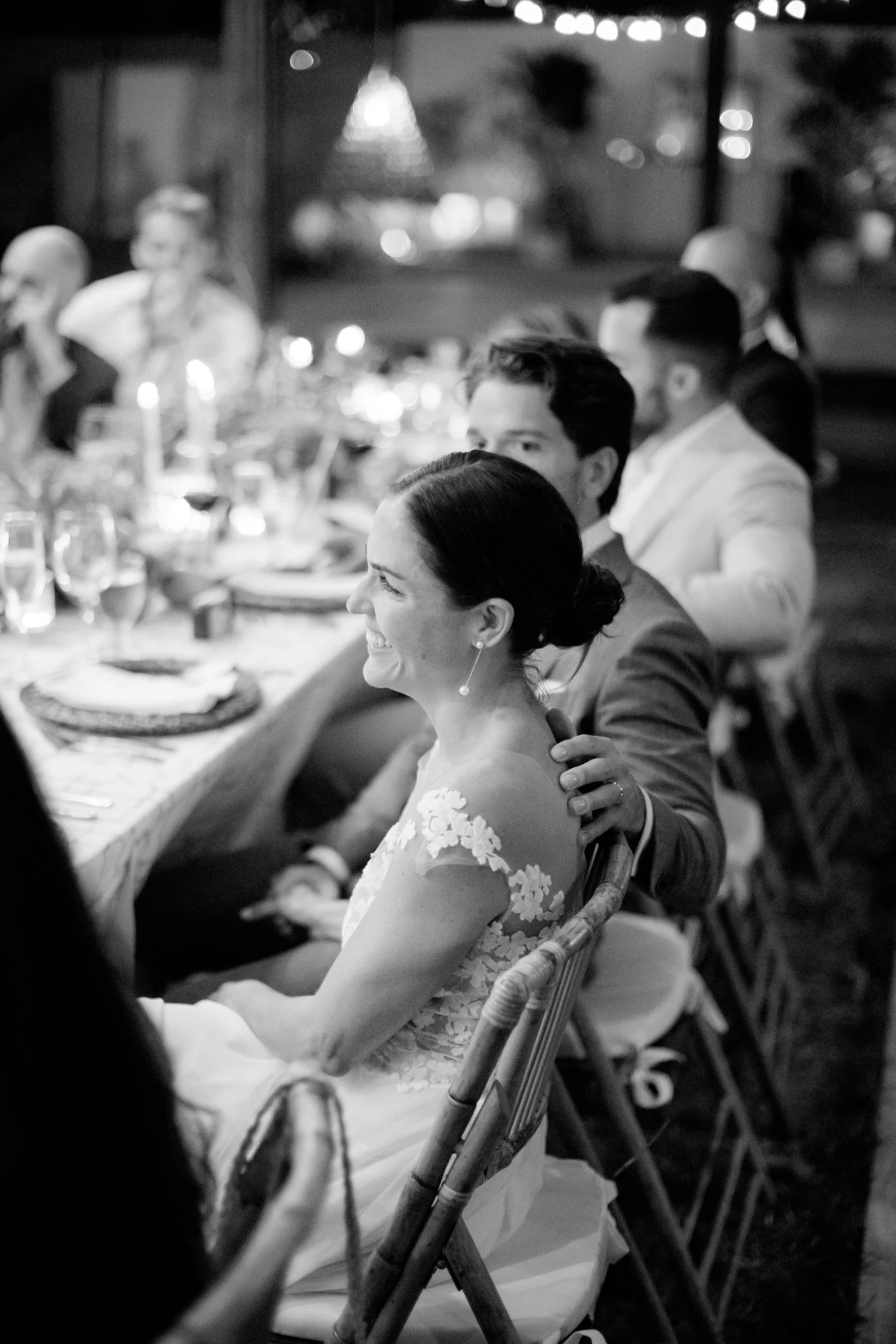 A woman in a lace dress smiling during a formal dinner, sitting at a long table with several men, all dressed in suits, in a decorated indoor setting.