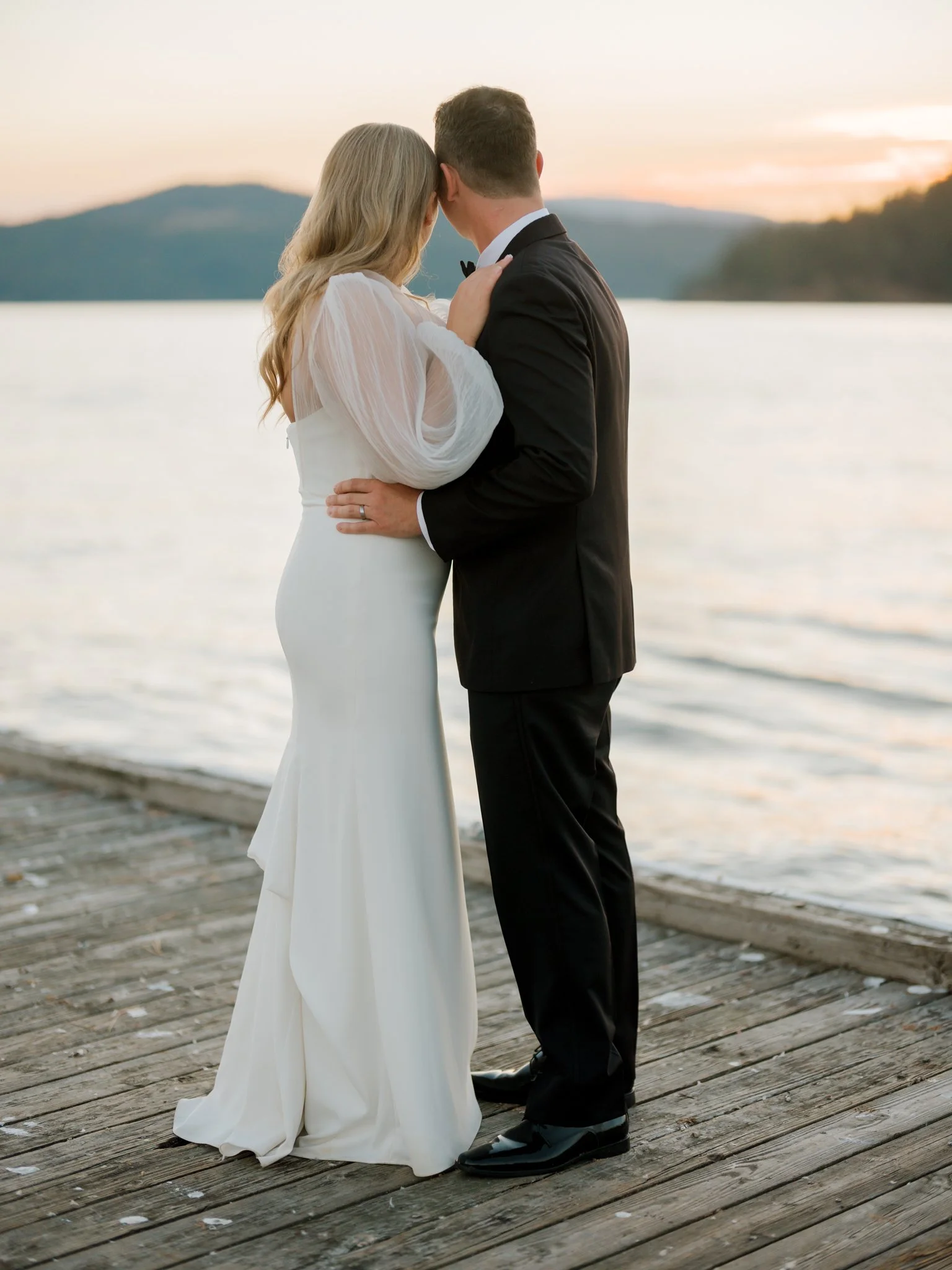 A bride and groom standing close on a wooden dock by a lake during sunset, with water, mountains, and a colorful sky in the background at the Hagadone Event Center in Coeur d'Alene, Idaho.