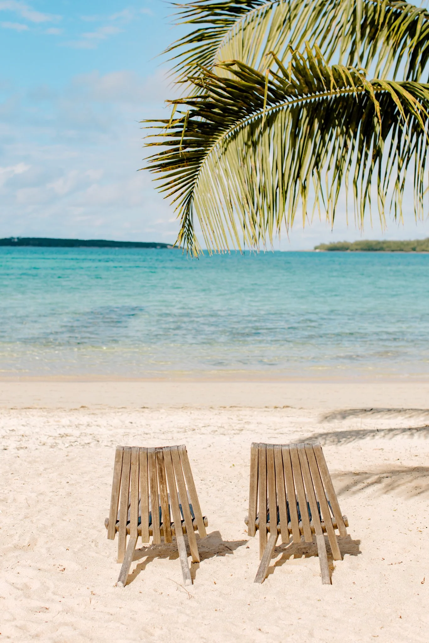 Two wooden lounge chairs on a sandy beach under a palm tree, with a view of turquoise ocean and blue sky.