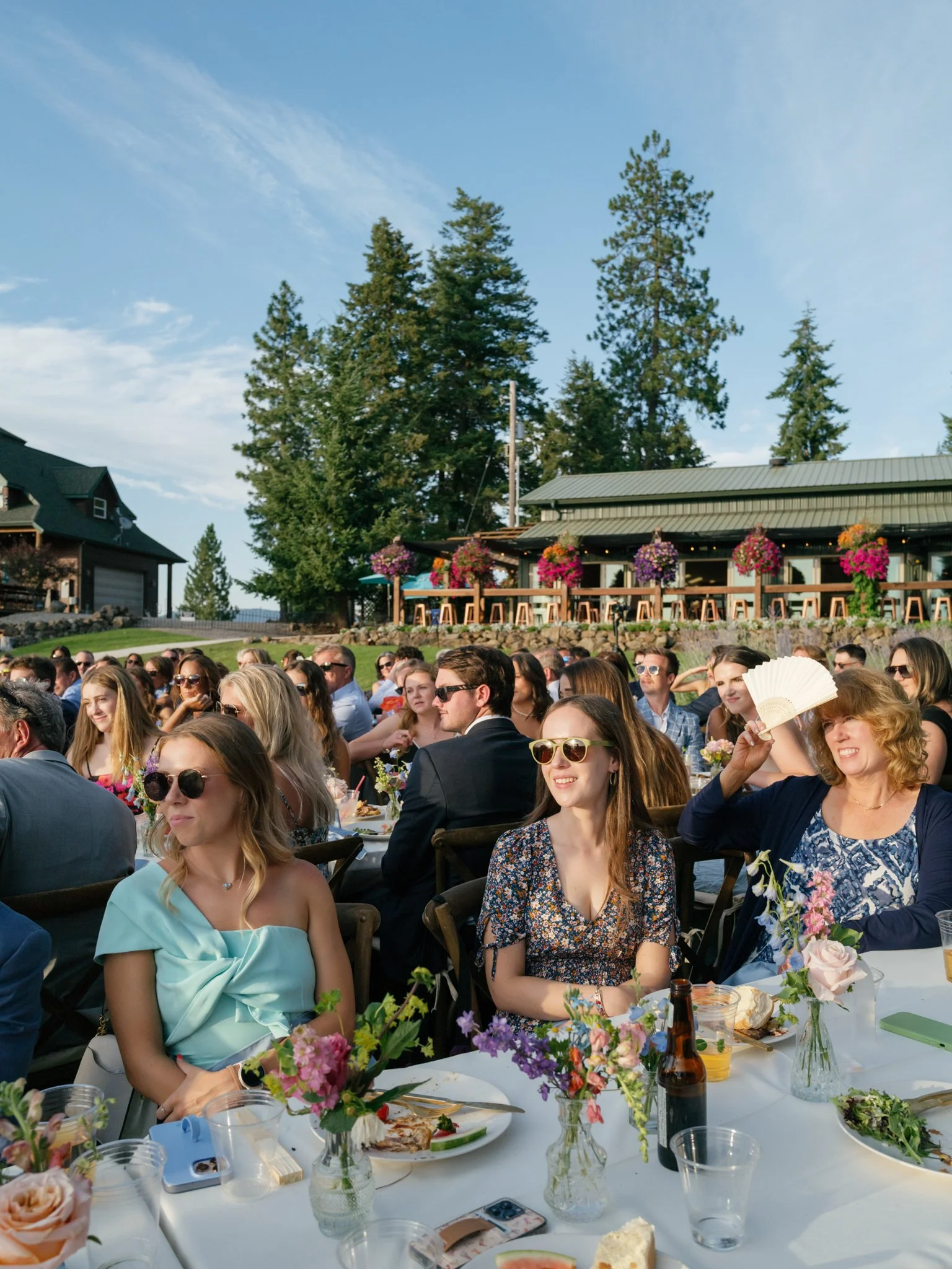 People attending an outdoor wedding reception, sitting at tables with floral centerpieces, enjoying food and drinks, surrounded by trees and a building decorated with hanging flower baskets at Elk Point wedding above Lake Coeur d’Alene.