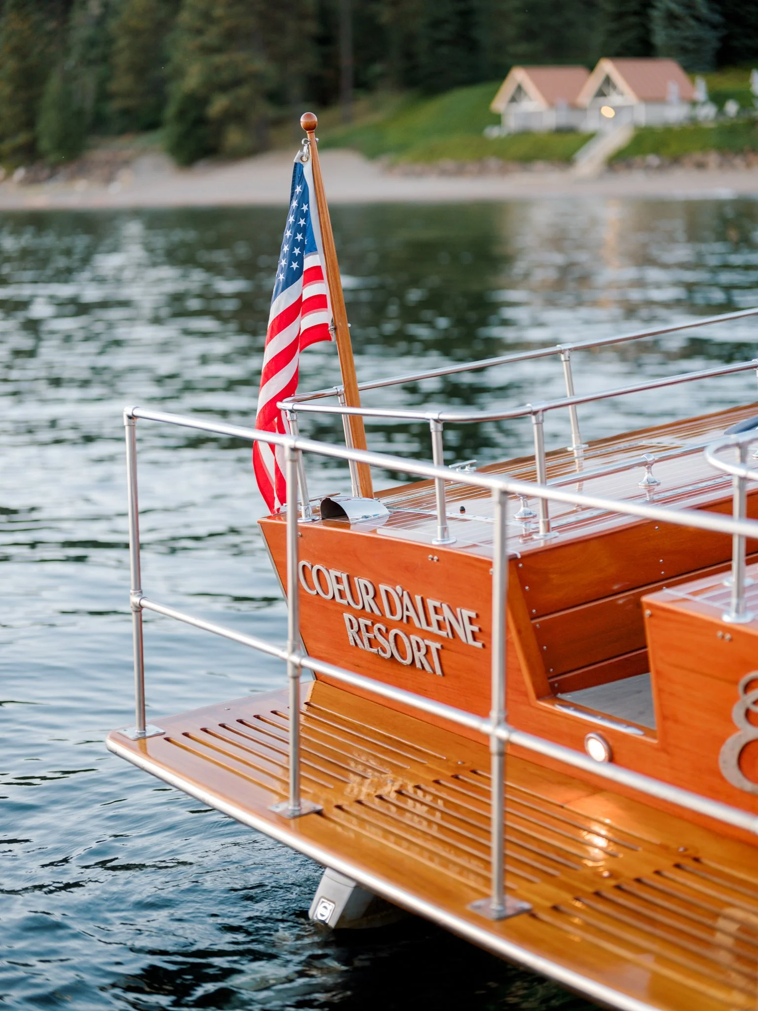 A wooden boat with the words 'Coeur d'Alene Resort' on its side, floating on a body of water with a house and trees in the background, and an American flag at the stern.