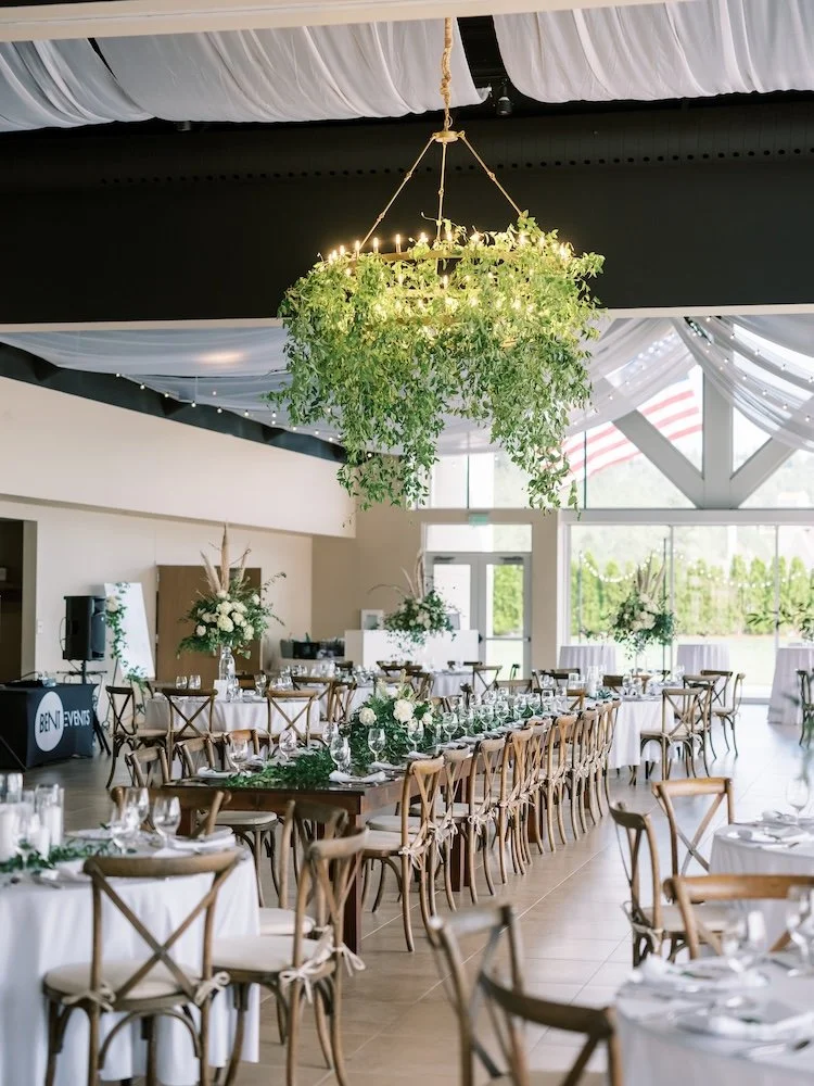Elegant event setup in a bright venue with greenery chandelier, long dining tables with white floral centerpieces, and wooden chairs with white cushions.