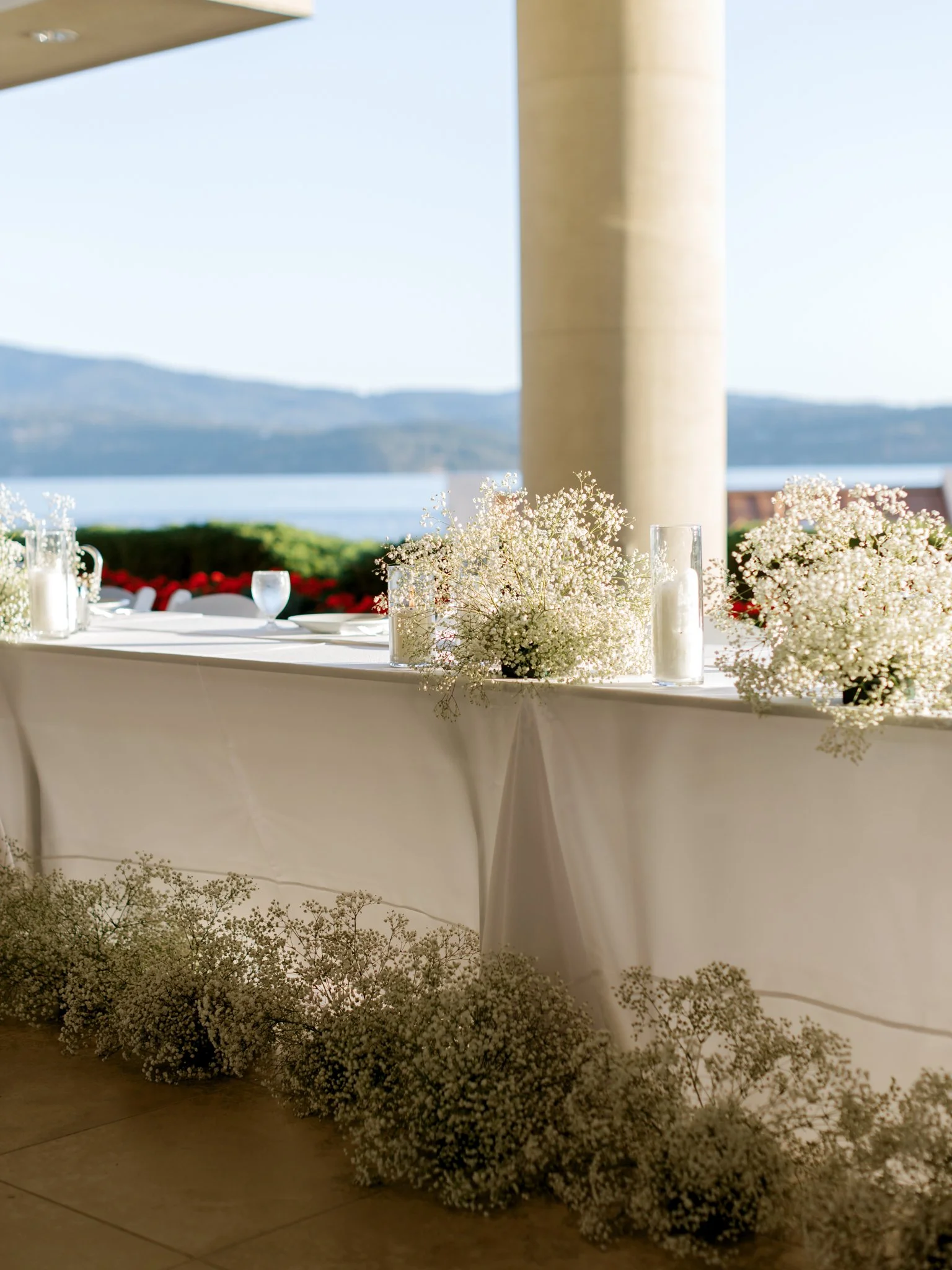A decorated table with white tablecloth, white floral arrangements, and candles, set outdoors with a view of water and mountains in the background.