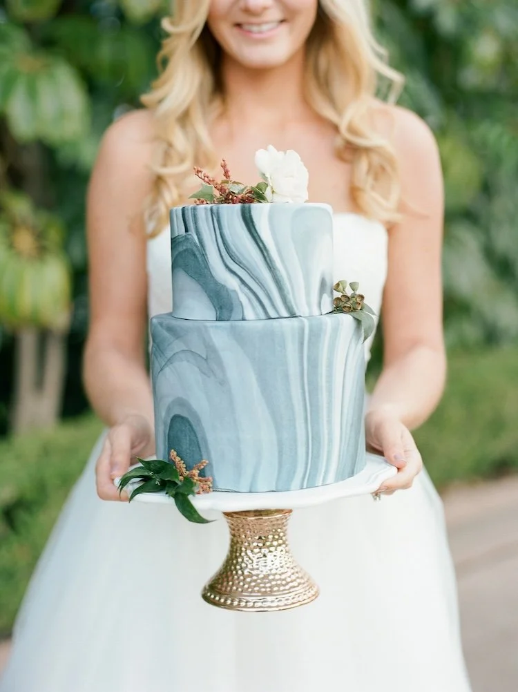 A woman in a white dress is holding a two-tier marble-patterned wedding cake decorated with small flowers and greenery, standing outdoors with blurred green foliage in the background.