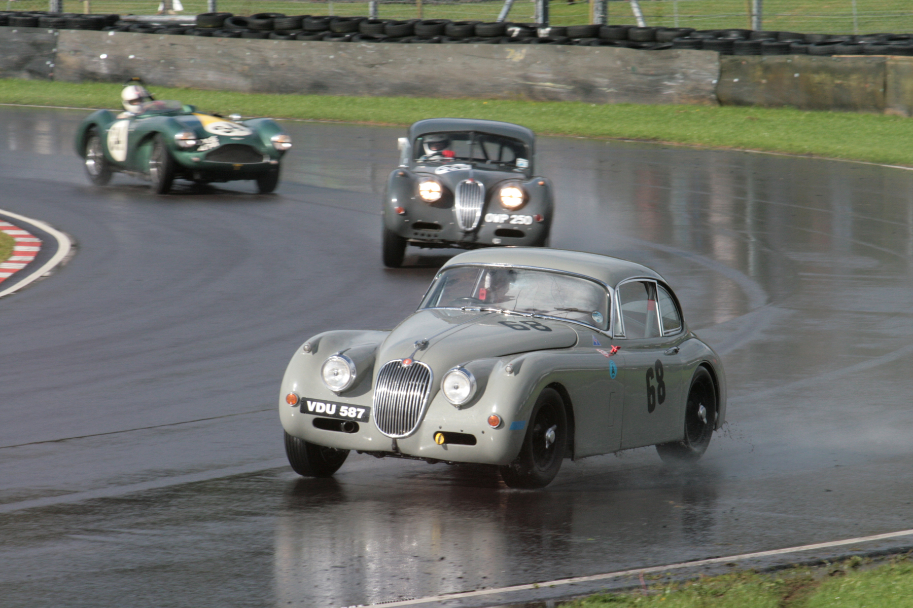Paul de Havilland takes a wide line out of Quarry in his Jaguar XK150 whilst looming large behind is the lead battle . Photo - John Turner