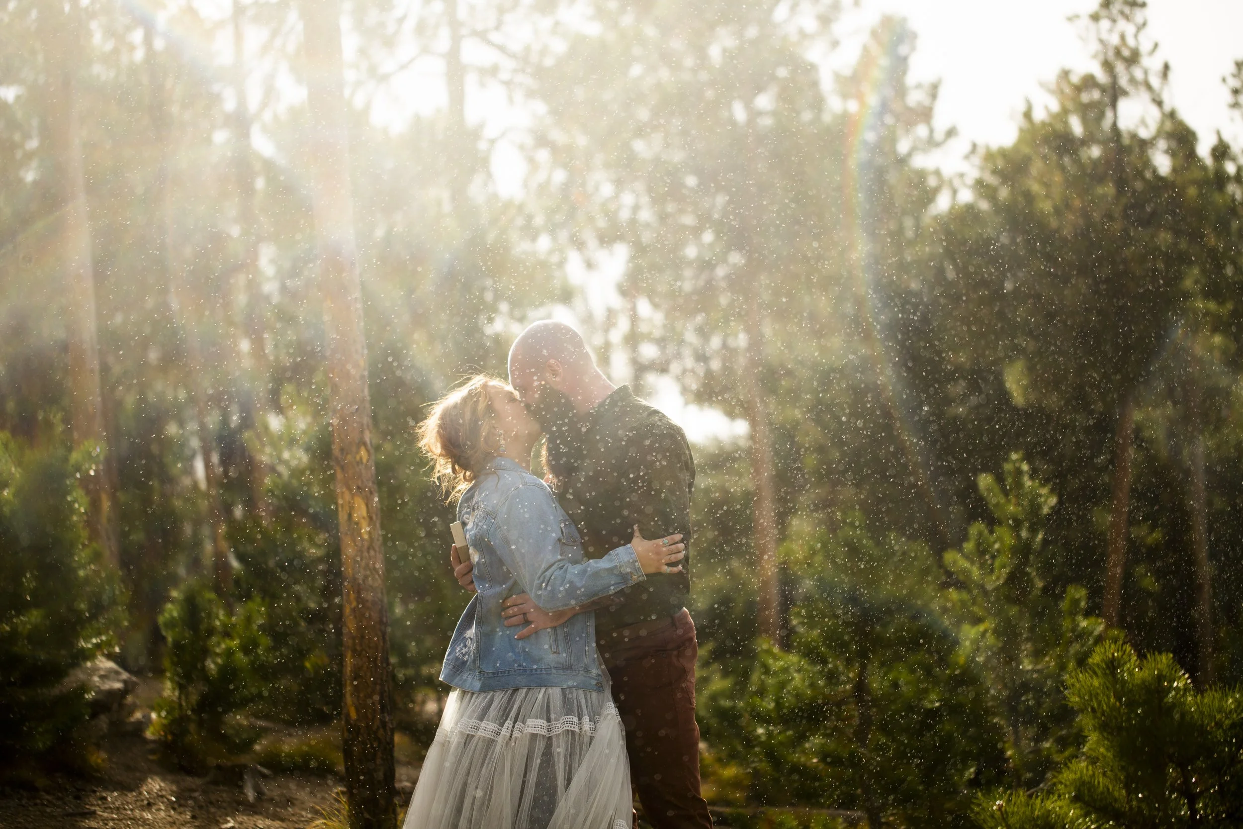 A couple sharing a kiss outdoors in a forest setting with sunlight and water droplets around them.