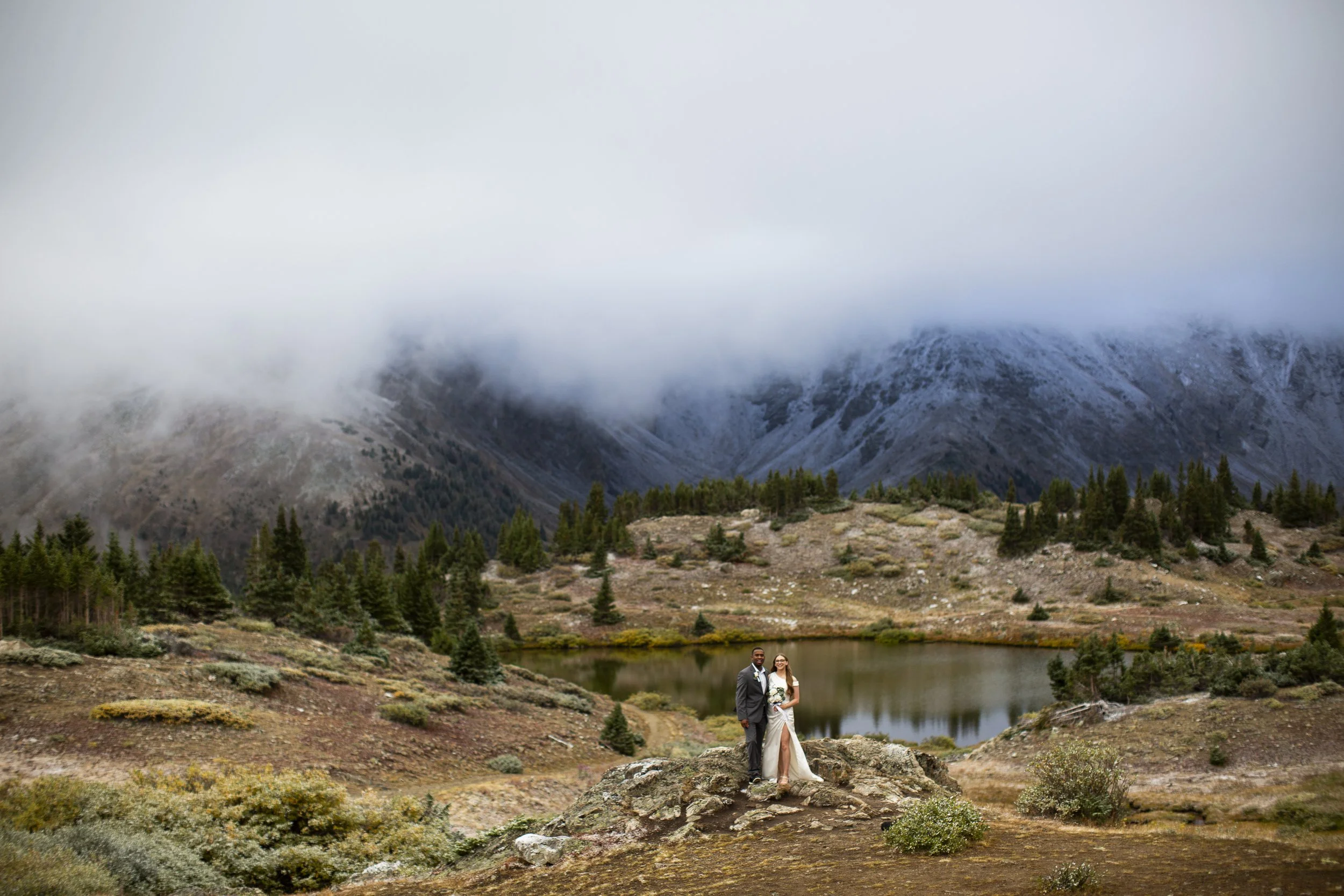 A bride and groom standing on a rock in a mountainous landscape with a lake, trees, and low-hanging clouds.