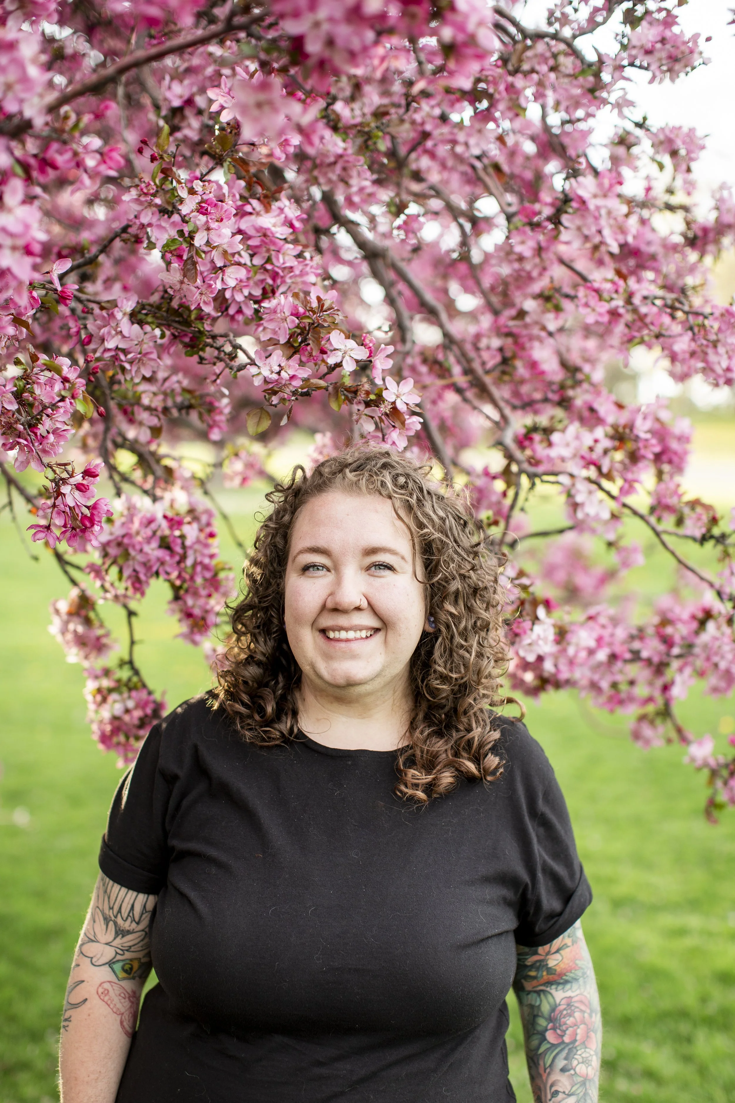 Young woman with curly hair smiling in front of a blooming pink tree on a sunny day.