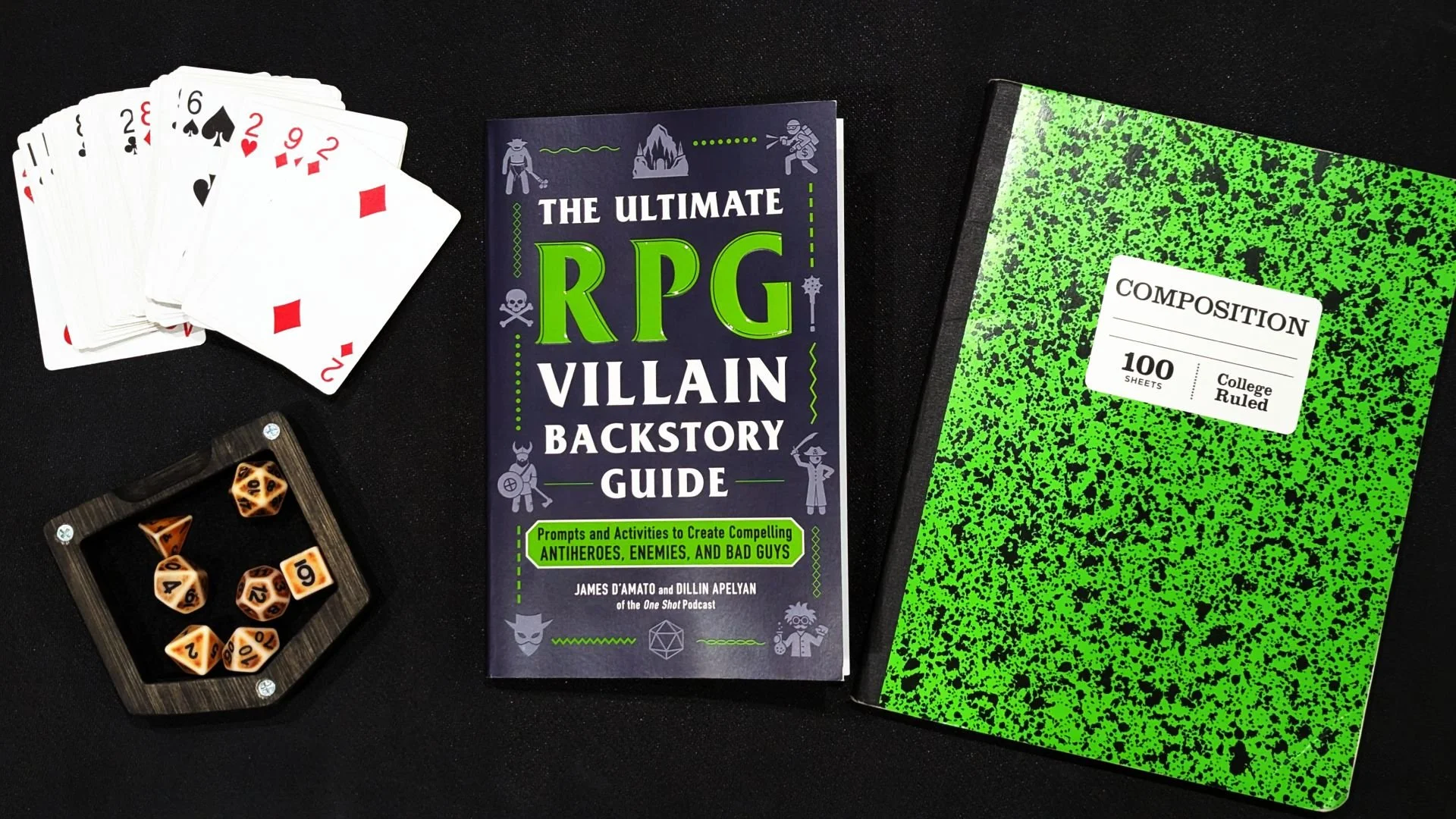 A book is in the middle with dice, a deck of cards, and a composition notebook around it.