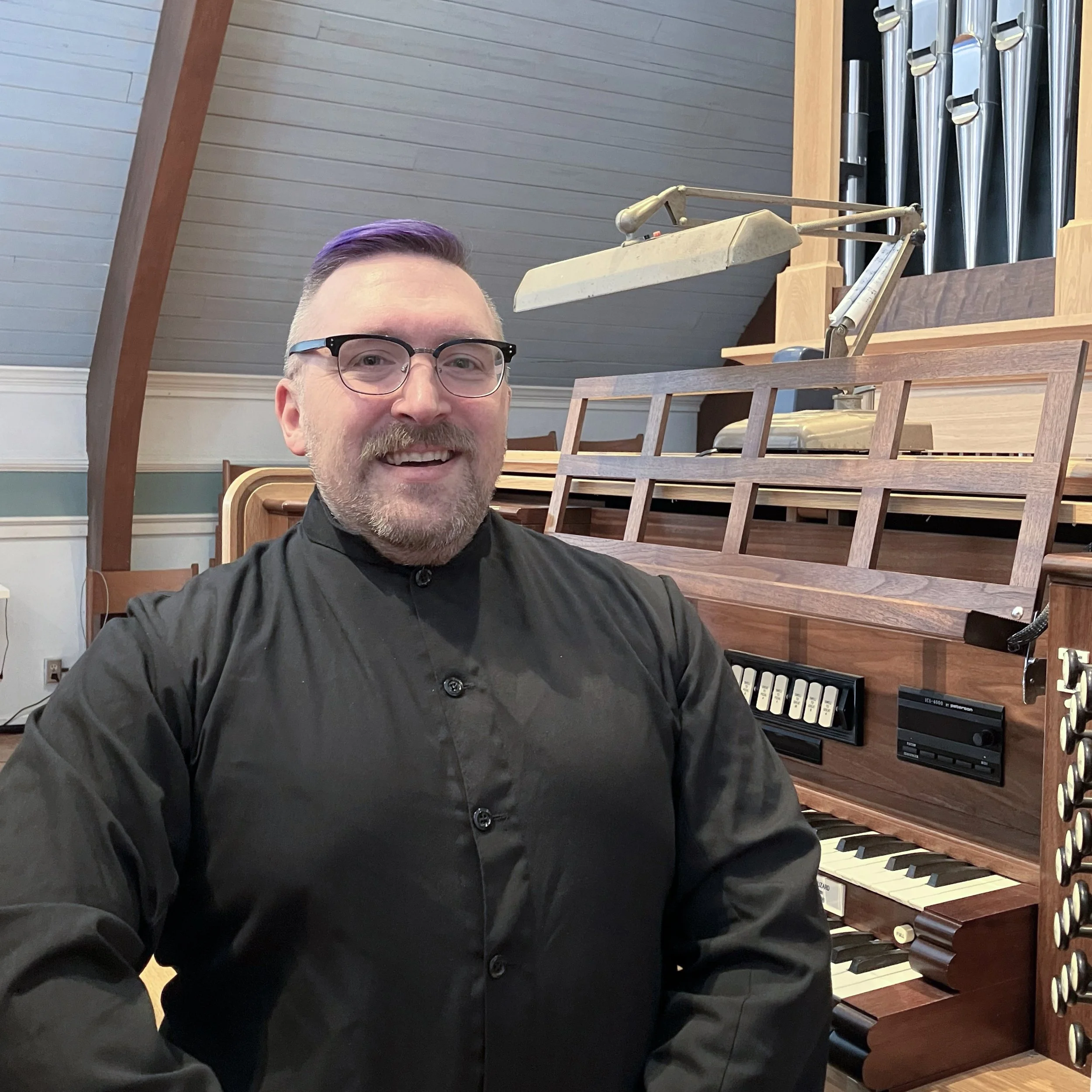 Robert sits at an organ bench – he has purple hair and glasses and is wearing a black cassock
