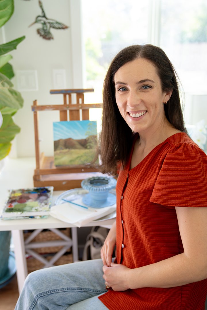 Portrait of artist Kristen Walker smiling in her Santa Barbara art studio, seated beside a tabletop easel with a Santa Cruz Island landscape painting and acrylic art supplies.