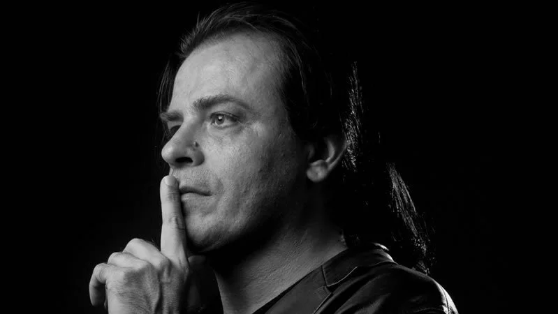 Black and white portrait of Tolga Katas with long hair, holding his index finger to his lips in a shushing gesture.