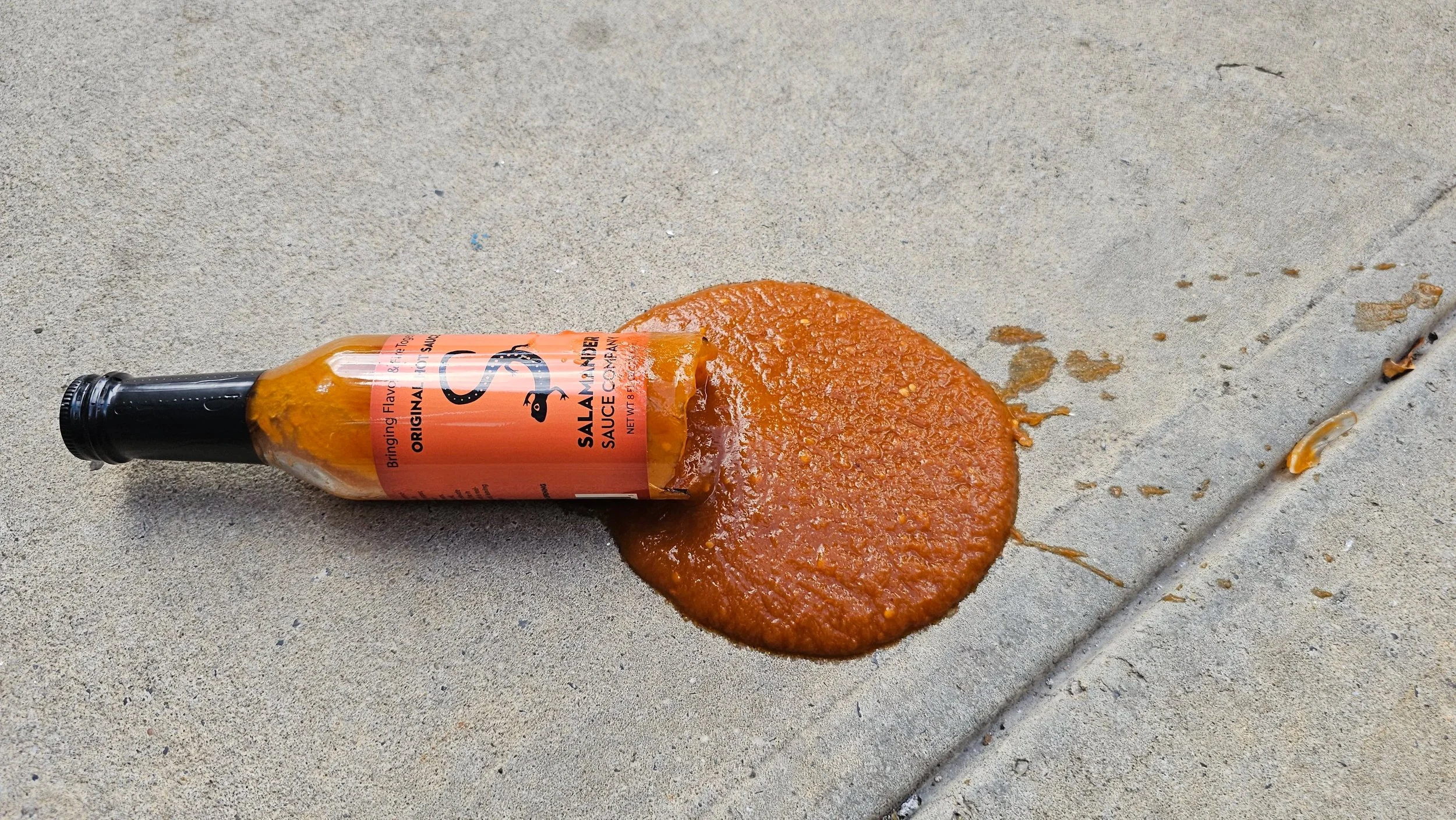 A broken bottle of Salamander Original on a concrete sidewalk, sauce pooled around it