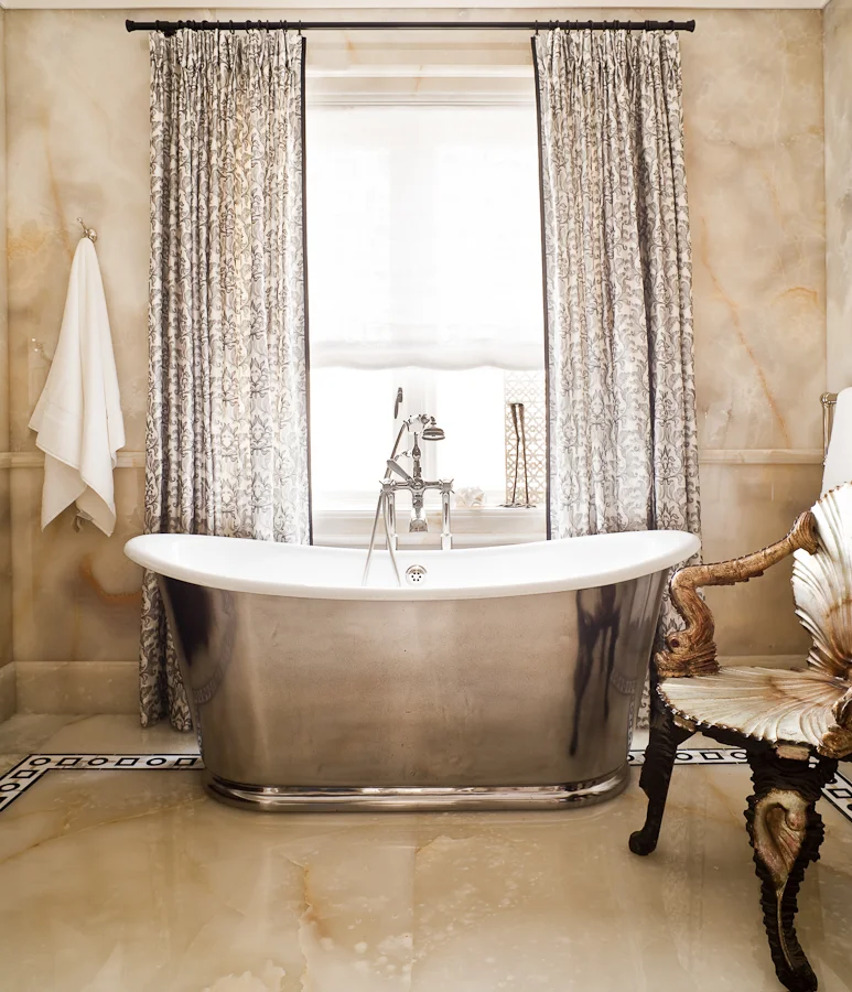 The Waterworks pewter tub and fixtures is the centrepiece of this bathroom. The pewter finish is a foil to set off the onyx walls and floors. One of my signature elements in my work is to add unexpected drama and warmth which here is achieved with the full length silk curtains and the antique shell chair. (Photography by Ted Yarwood) 