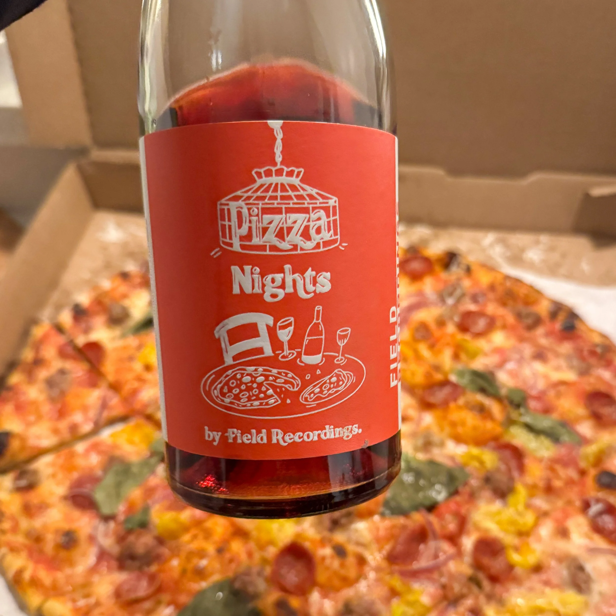 A bottle of Pizza Nights wine, which has a red label and an illustration of a pizza dinner, held in front of a pizza in a cardboard box.