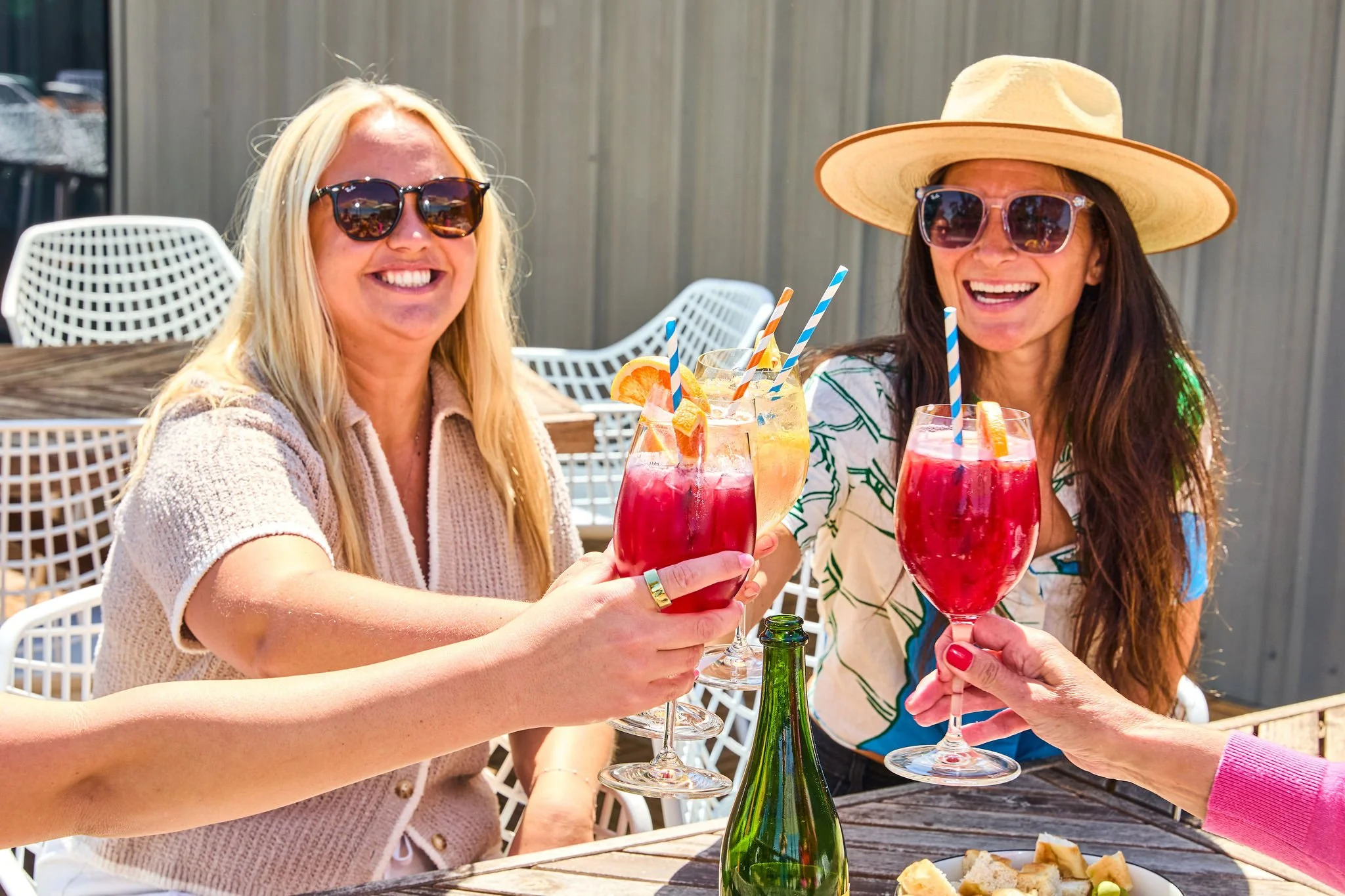 Two women celebrating outdoors with colorful cocktails garnished with fruit slices and striped straws, smiling and wearing summer accessories like large sunglasses and a hat.