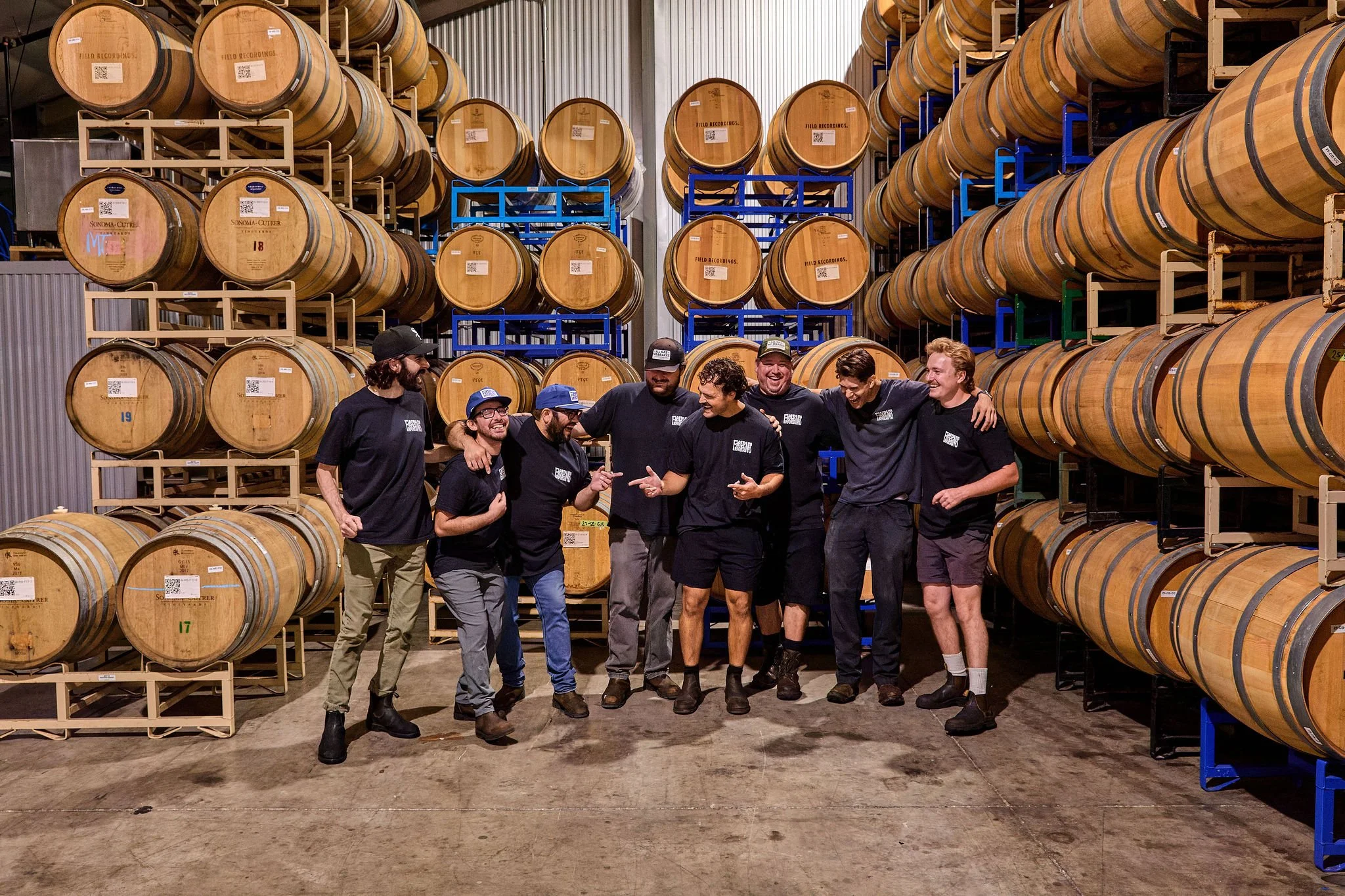 A group of cellar workers smiling and laughing with each other, in front of barrels in the Field Recordings wine cellar.