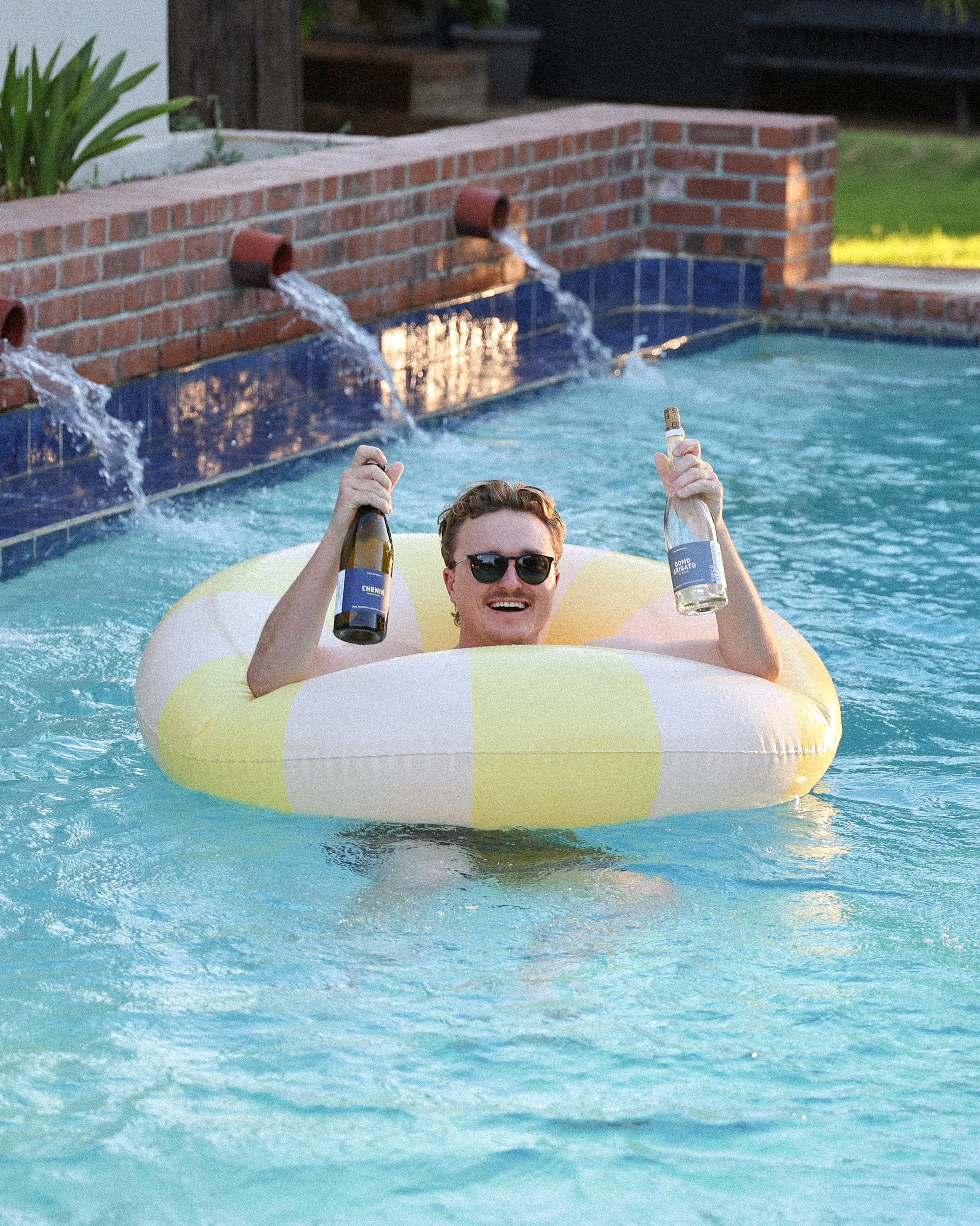 Jack floats in a white and yellow floatie in the pool. He's holding up two bottles of wine.