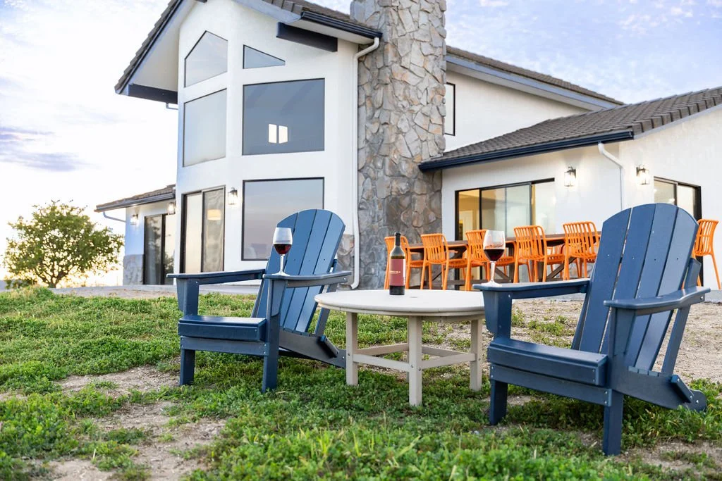 A house with large windows and a stone chimney, outdoor seating with two blue Adirondack chairs, a small white table with wine and a bottle, and a dining table with orange chairs.