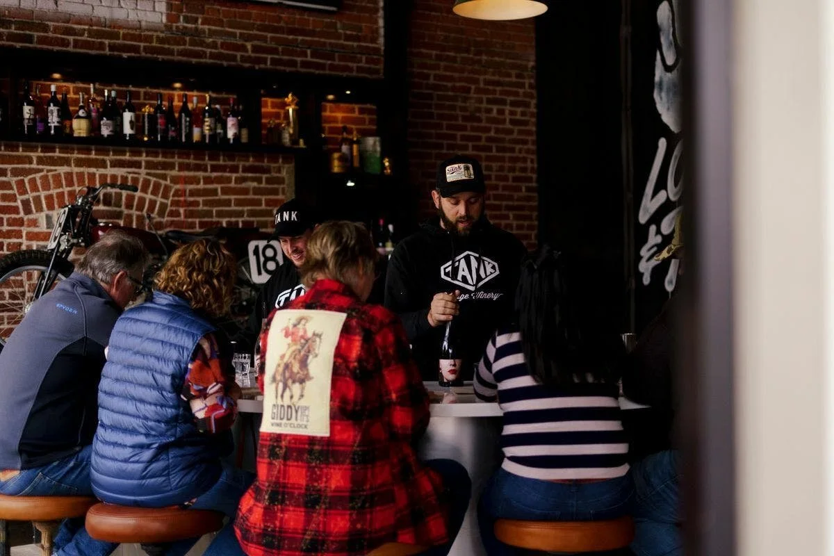 Three wine tasters sit at a bar at Tank Garage Winery. Two men wearing black sweatshirts and hats stand behind the bar.
