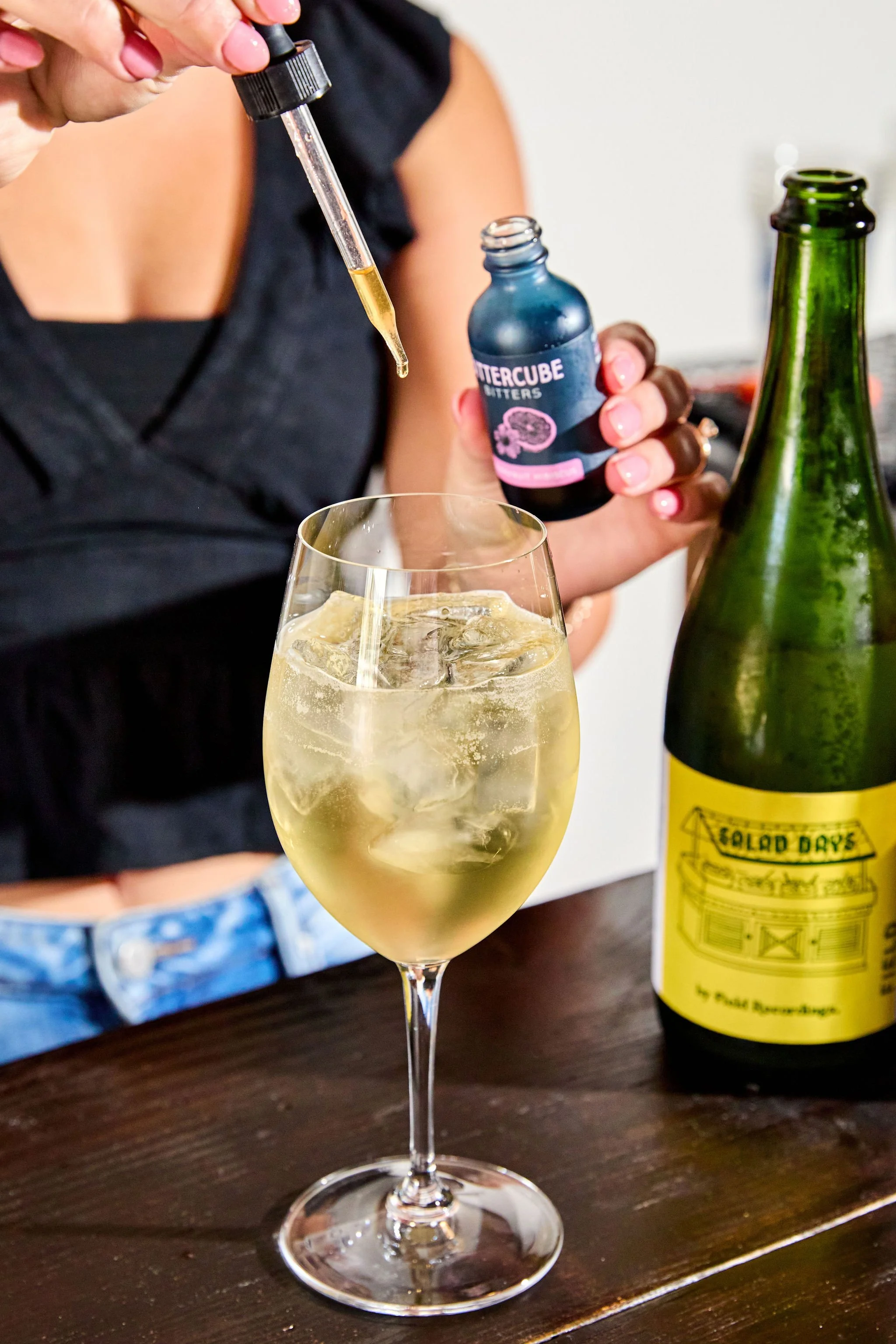 A person is using a dropper to add bitters into a spritz cocktail made with Field Recordings sparkling wine. The glass is on a dark wooden table.