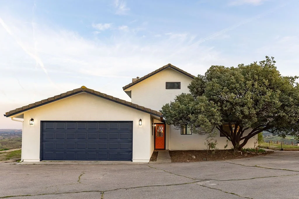 A modern house with a dark gray garage door, white exterior walls, a red front door, and a large tree in front, set against a blue sky.