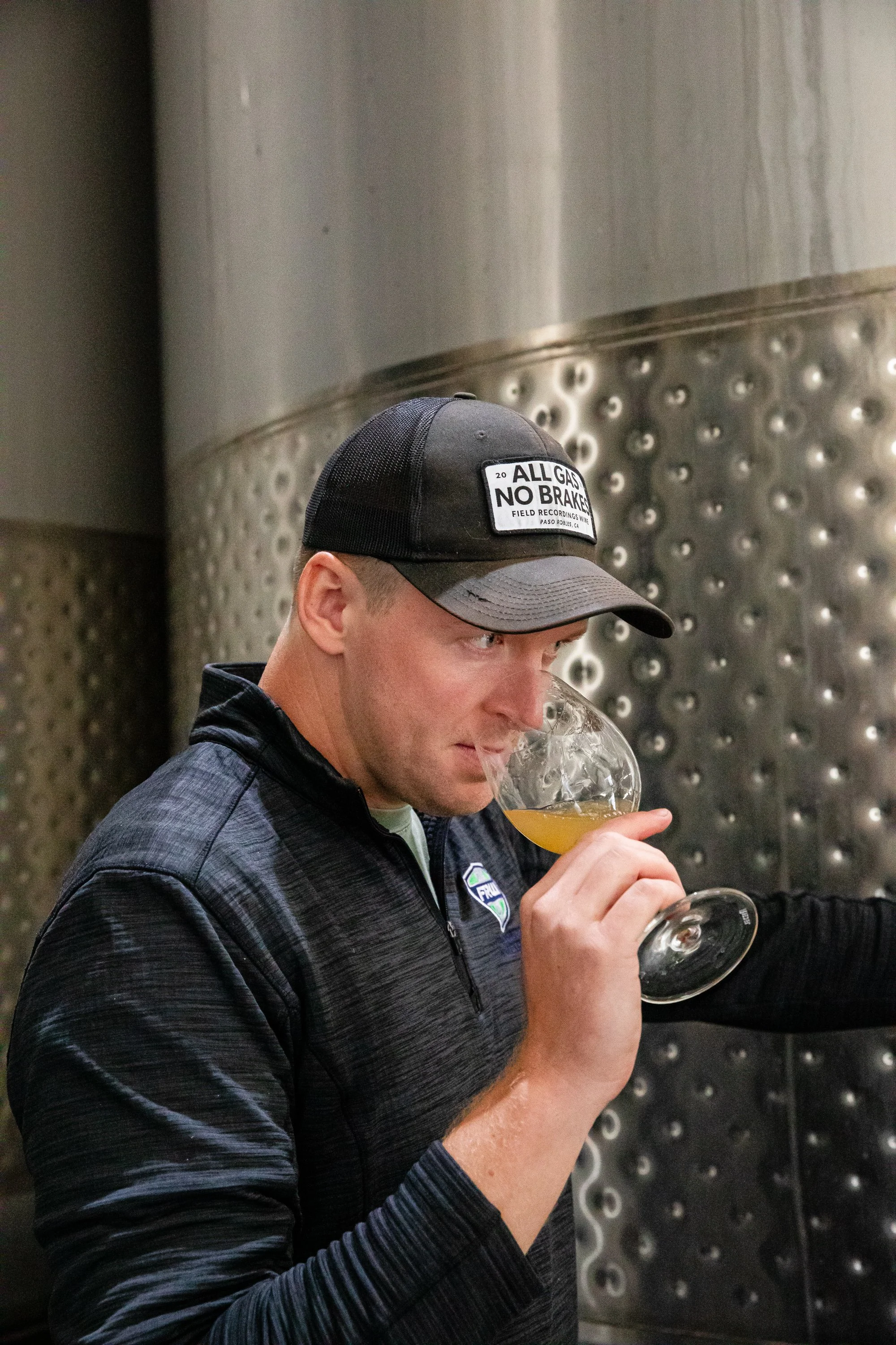 Tim, wearing a black sweater and black hat, smells a glass of white wine juice in front of stainless steel tanks.
