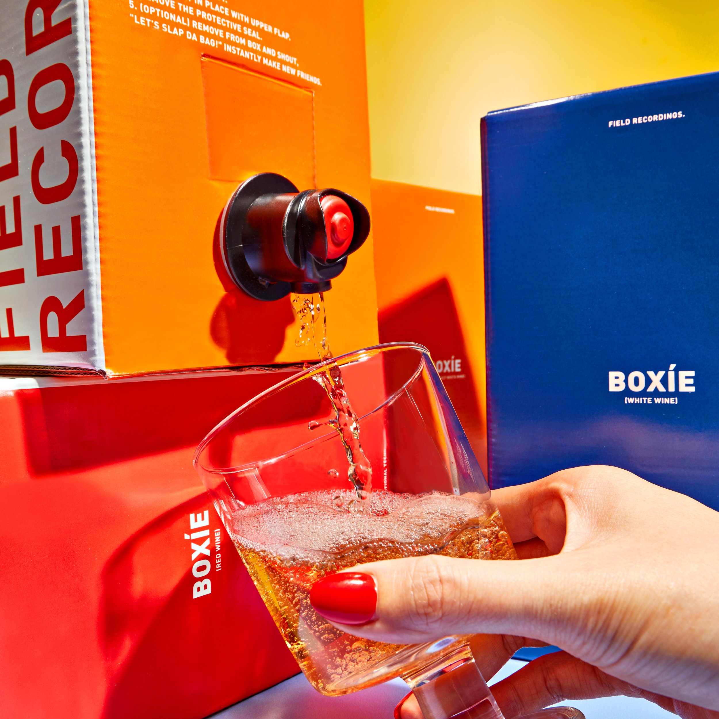 A hand holds a glass of wine that's being filled from a boxed wine spout. There are boxed wines stacked around.