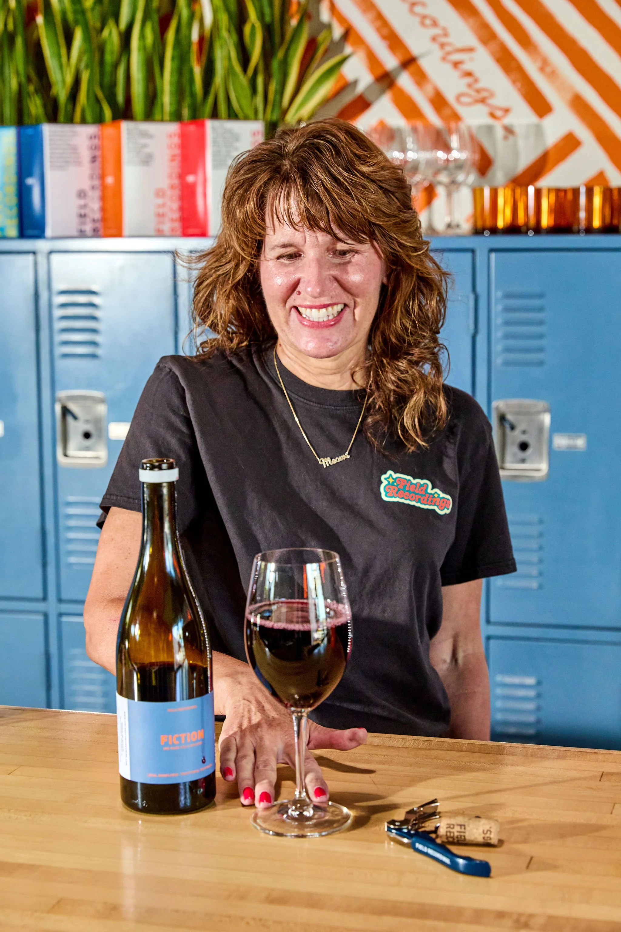 A woman with red hair and a black shirt that says 'Field Recording' sitting at a table with a wine glass, wine bottle, corkscrew, and corks in the foreground. She is smiling and has red nail polish.